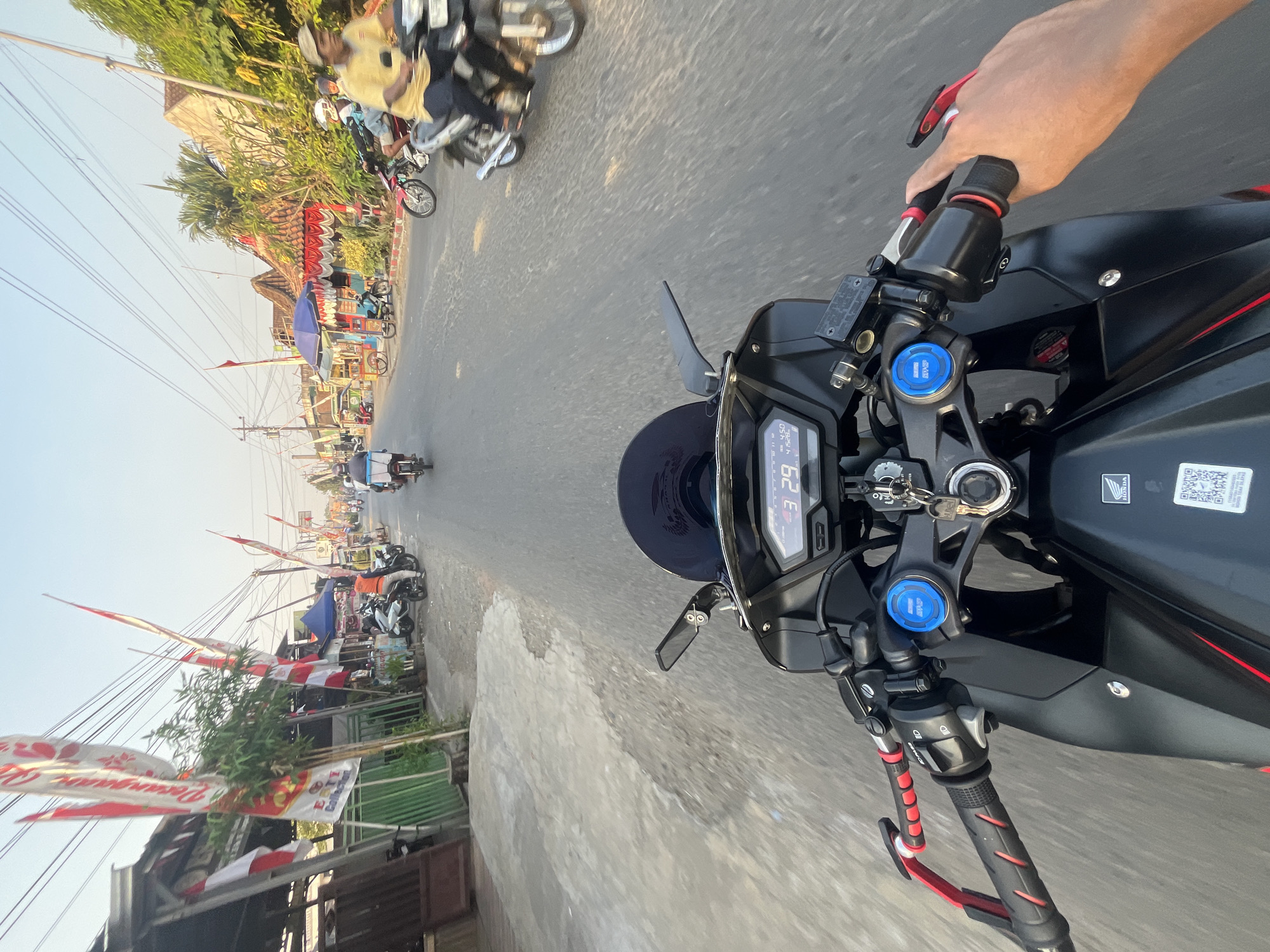 This is a first-person perspective, point-of-view shot from a motorcycle traveling on a paved road in Pecangaan, Indonesia. The road is lined with buildings and trees, and there are numerous red and white Indonesian flags displayed, suggesting a national celebration. Several motorcycles and a few other vehicles are visible on the road and the side. The sky is clear and bright, indicating daytime. The foreground of the image is dominated by the motorcycle's handlebars and dashboard, displaying a digital speedometer reading "43 29". A human hand is visible on the right handlebar.