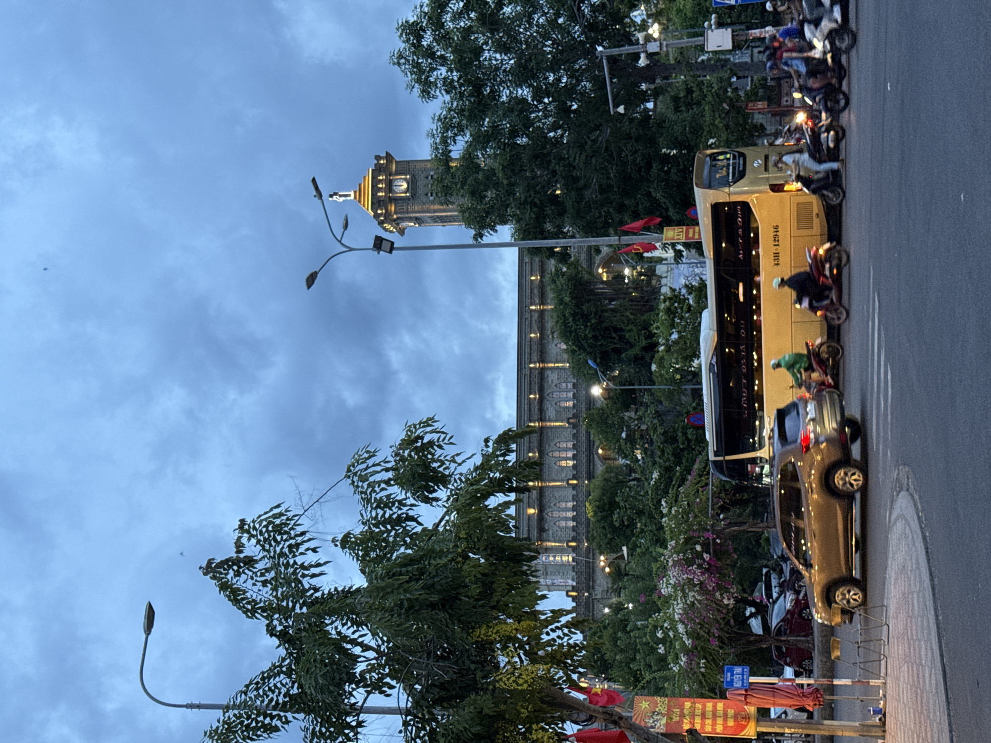 In Nha Trang, Vietnam, an urban street scene unfolds under a cloudy, twilight sky. A prominent, stone-clad building with illuminated windows and decorative lighting forms the backdrop. A clock tower, topped with a statue and illuminated, rises from the building's structure, serving as a focal point. Trees with lush green foliage and flowering bushes line the street and surround the building, with some branches extending into the foreground.
The street is active with traffic. A large, gold-colored bus with illuminated lettering drives on the right side of the road. A gold SUV is driving on the left side of the road, turning. Several motorcycles, each carrying one or two individuals, are also present, some driving alongside the bus and SUV, while others are stopped or in motion. People are visible on the motorcycles, some wearing helmets.
Streetlights, including one with multiple lamps on a tall pole, are present, indicating the transition from day to night. Red flags and a banner with text are attached to a pole on the left side of the image. The road surface appears to be asphalt, with a pedestrian walkway in the foreground. The overall atmosphere suggests a busy urban environment during the evening hours.