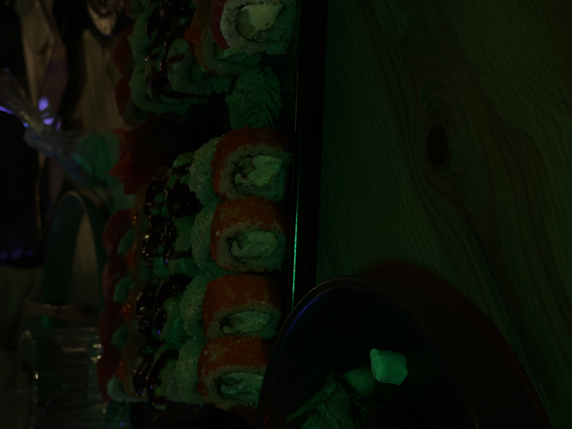The image shows a close-up of sushi rolls on a platter next to a wooden surface, possibly a wall. The sushi rolls have a variety of toppings, including what appears to be roe or a similar orange-colored ingredient. A black bowl containing what could be wasabi or another condiment is partially visible in the foreground. The lighting is dark with a greenish hue, obscuring fine details. There's insufficient information to determine the exact location in Yerevan, Armenia, without additional context or clues. No people are visible in the image.