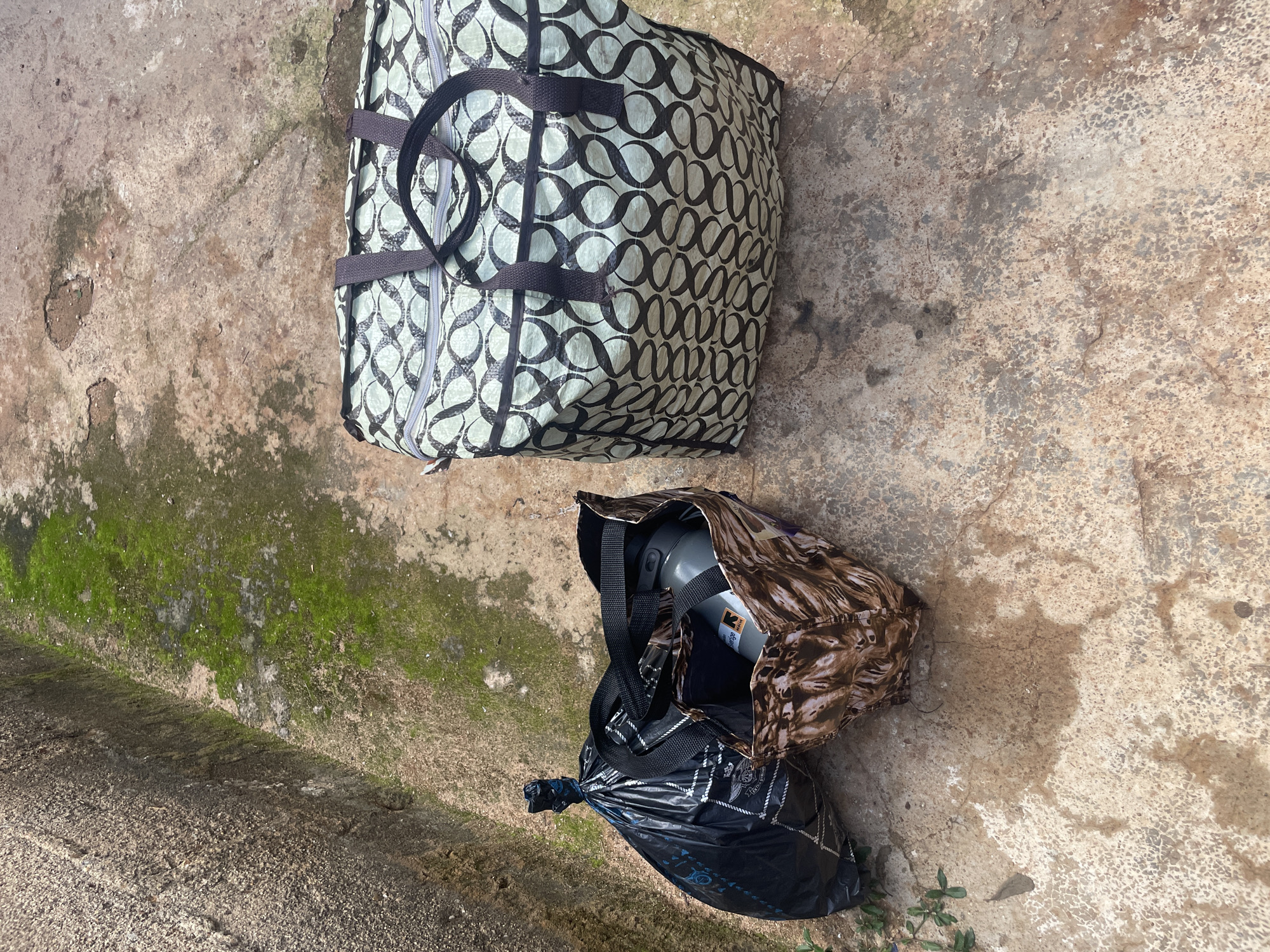 The image shows three bags placed on a concrete surface. A large black trash bag, tied at the top, is on the left. To its right is a brown patterned shopping bag, which contains a gray water bottle. To the right of these two bags is a large, light green and black patterned tote bag with handles. The background consists of a moss-covered concrete wall on the left and a textured concrete ground with some patches of moisture. The location is identified as Udu, Nigeria.