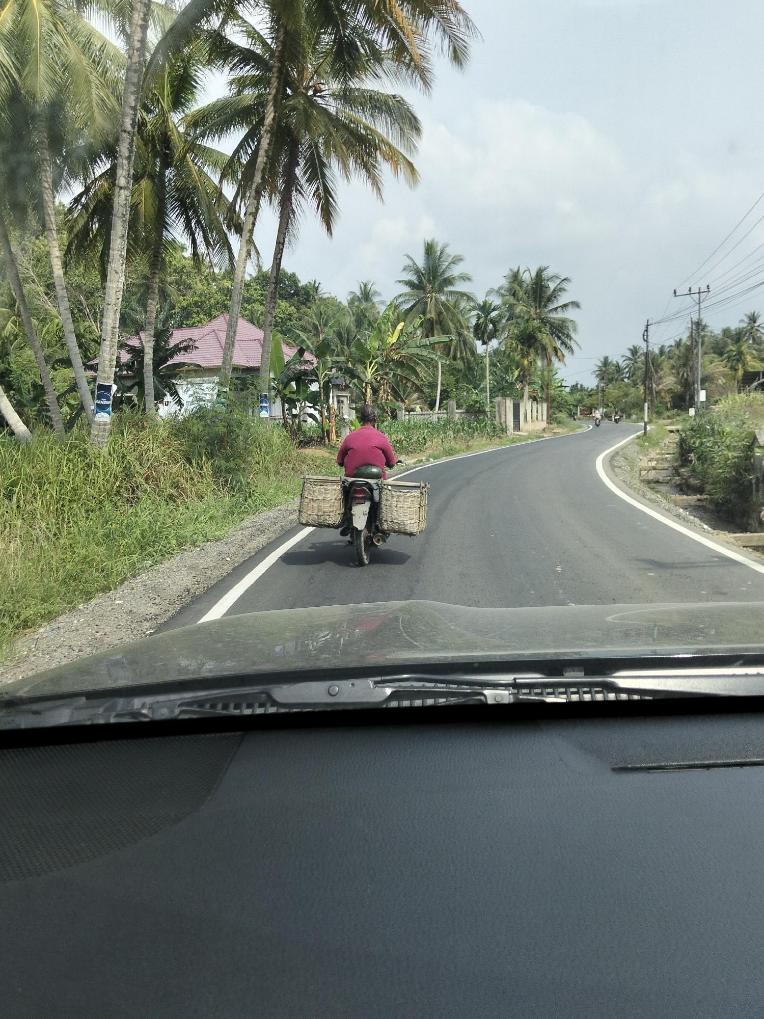 A person rides a motorcycle with two large woven baskets attached to the rear in Luengbaro, Indonesia. The person is wearing a reddish-pink shirt. The motorcycle is traveling on an asphalt road with a white line marking the lane. The road curves to the right. On the left side of the road, there is a grassy shoulder and several tall palm trees, with a house visible in the background. On the right side of the road, there are more trees and vegetation, as well as a utility pole with wires. The sky is overcast with some clouds. The image appears to be taken from inside a vehicle, with the dashboard and windshield wipers visible in the foreground.