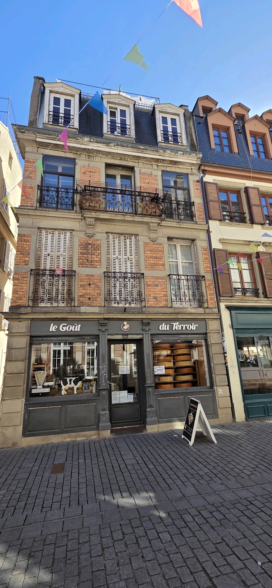 The image shows the exterior of a cheese shop called "Le Goût du Terroir" located on a cobblestone street in Strasbourg, France. The shop is situated on the ground floor of a multi-story building with French-style architecture. The facade features brick and stone elements, with multiple windows on the upper floors and decorative iron balconies.
The shop's storefront has large display windows showcasing various cheeses, including large wheels of cheese stacked in the right window. Inside the left window, a black and white cow figurine stands next to a vintage scale. The shop's name, "Le Goût du Terroir," is prominently displayed above the entrance. A sign with a drawing of cheese and text in French is placed on an A-frame stand outside the shop.
Colorful triangular flags are strung across the blue sky above the building, suggesting a festive occasion or decoration. The street is paved with dark cobblestones, and sunlight casts shadows on the ground. The building to the right of the cheese shop also has shutters and windows typical of Strasbourg's architecture. The overall scene depicts a charming street view in a European city.