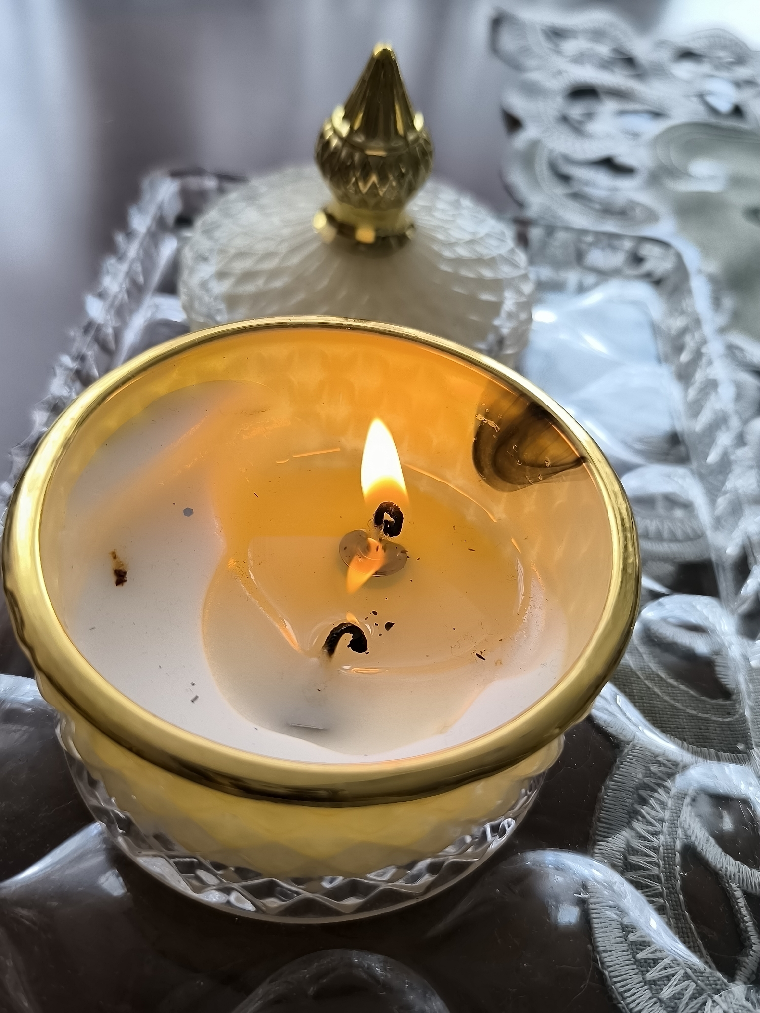 The image is a close-up shot of a lit candle in a decorative glass and gold holder. The candle appears to be partially melted, with a golden liquid wax pool around the wick. The flame is bright yellow and flickers slightly. In the background, out of focus, is another decorative object with a gold finial and a textured white surface. The candle is placed on a clear glass tray with a decorative edge. The tray itself rests on what appears to be dark, polished stones or glass. A textured, white lace-like material is visible beneath and to the right of the tray. The lighting is warm and focused on the candle, creating a soft glow. There are no people or any identifiable landmarks that would indicate a specific location in Klaipėda, Lithuania.