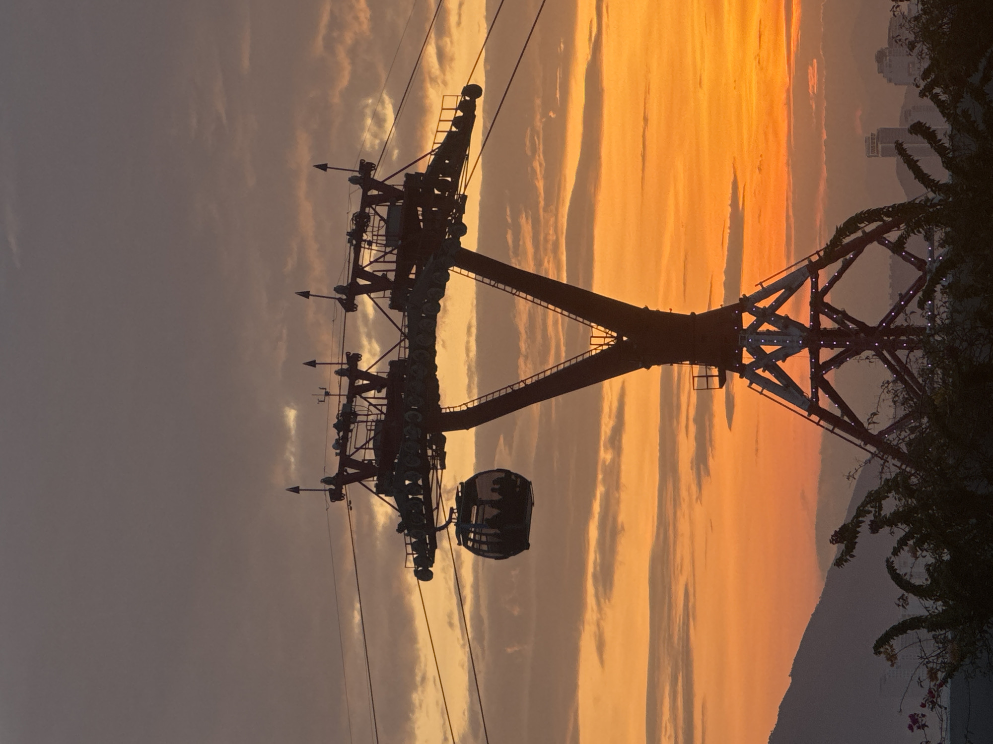 A cable car station with a cable car attached to it is silhouetted against a vibrant orange and purple sunset sky over the city of Nha Trang, Vietnam. The cable car station is a tall, metallic structure with a V-shaped support. The cable car is a rounded, glass-enclosed gondola, and it appears to be carrying passengers. In the background, silhouetted mountains and city buildings are visible. The foreground is filled with dark foliage and branches. The overall mood of the image is serene and picturesque, capturing the beauty of a sunset over a coastal city.