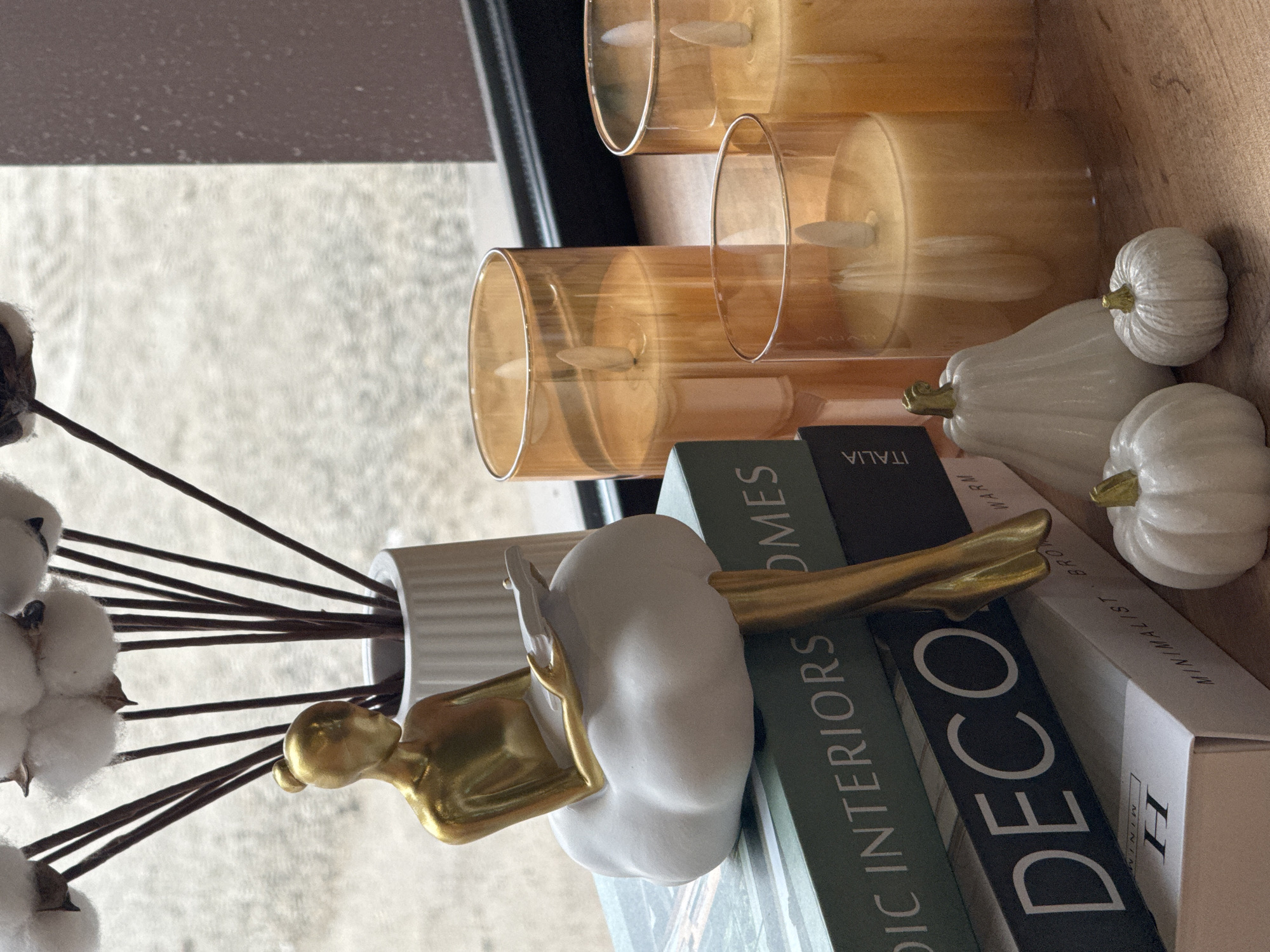 The image depicts a still life arrangement. Three cylindrical amber glass candle holders containing artificial candles are visible. Three small, white decorative pumpkins are positioned near the candles. A white vase holding cotton boll stems sits near a decorative figurine of a person with a white body and gold details. The figurine rests on stacked books with titles related to interior design. The location is not discernible from the image.