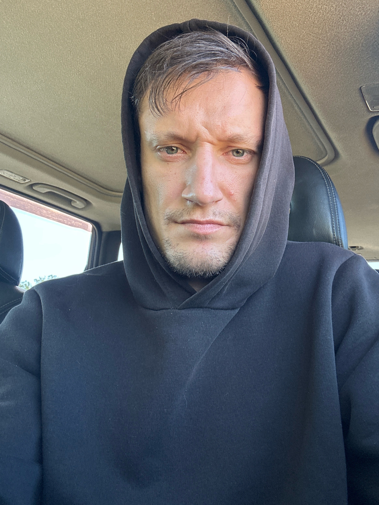 The image is a close-up, selfie-style portrait of a man wearing a dark hooded sweatshirt, with the hood pulled up. He is seated in the passenger seat of a car, as evidenced by the car's interior visible in the background, including the car's ceiling and part of a seat. The man has light-colored eyes, short brown hair peeking out from under the hood, and a light beard stubble. His facial expression appears concerned or questioning, with furrowed brows. The lighting suggests it is daytime, possibly sunny, with light coming from the right side of the frame. The image does not provide any explicit information about the specific location within the city of Makiv, Ukraine, but depicts a person in a car.