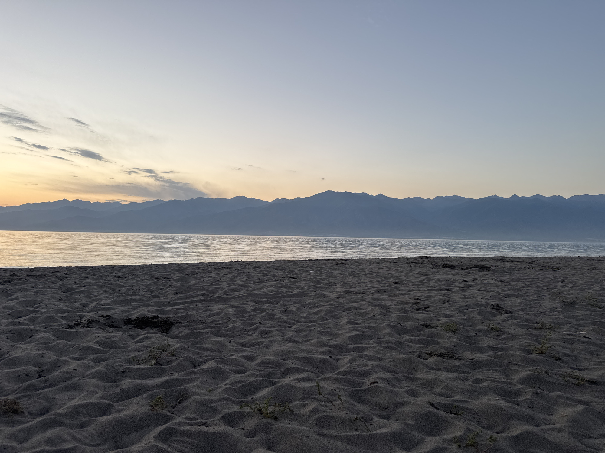 A wide shot shows a dark, rippled sand beach in the foreground. In the middle ground, a calm, gray body of water stretches horizontally, reflecting the sky. In the background, a long, undulating mountain range is silhouetted against a pale, gradient sky that transitions from a soft orange near the horizon to a light blue at the top. The scene appears to be a natural landscape, likely a lakefront with mountains in the distance, characteristic of the environs of Priozernoe, Kyrgyzstan. There are no people or man-made objects visible in the frame.