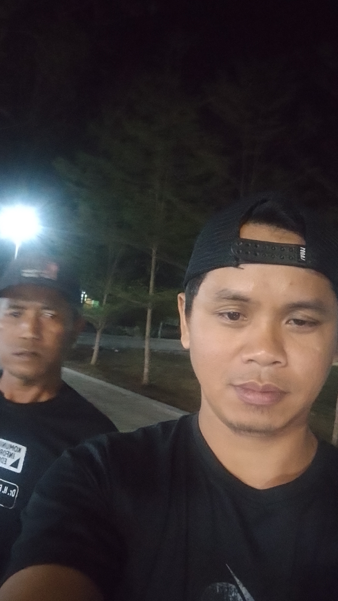 Two men are in a selfie taken at night. The man in the foreground on the right is wearing a black t-shirt and a black baseball cap. The man in the background on the left is wearing a black t-shirt and a black baseball cap. They appear to be outdoors, with trees and streetlights visible in the background. The location is identified as Karanglangu, Indonesia.