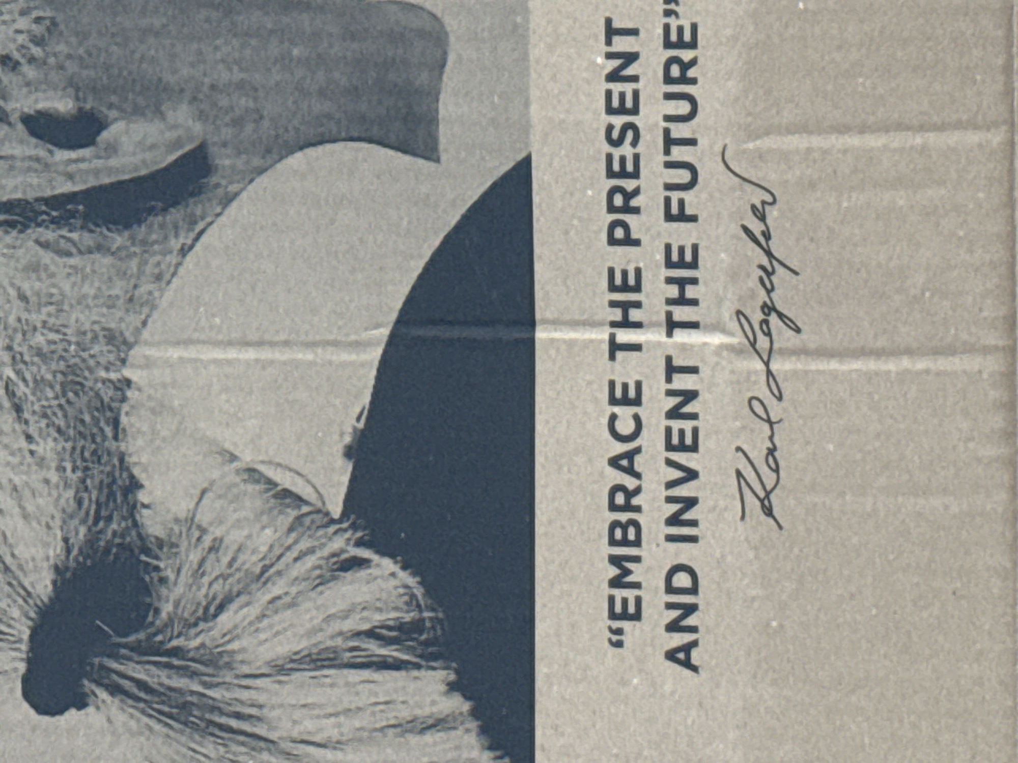 The image displays a stylized, predominantly monochromatic depiction printed on a textured surface, possibly cardboard, with a visible horizontal fold line across its center.
On the left side, a graphic illustration of a head or bust is visible. It features a dark, possibly silhouetted, profile of a face and neck, topped with a striking, voluminous white wig or elaborate hairstyle that appears fluffy and highly textured. The background behind this figure is a mottled dark gray.
The right half of the image is a lighter, plain gray-brown background. Vertically oriented text is printed in dark letters on this side. The text consists of a quote in all capital letters: "EMBRACE THE PRESENT AND INVENT THE FUTURE". Below the quote, a handwritten signature is present, clearly identifiable as "Karl Lagerfeld".
The overall aesthetic is simple and bold, with a strong contrast between the dark elements of the figure and text against the lighter background. The grainy texture throughout suggests a print on a coarse material. No specific time of day, weather, or location cues are discernible from the image itself.