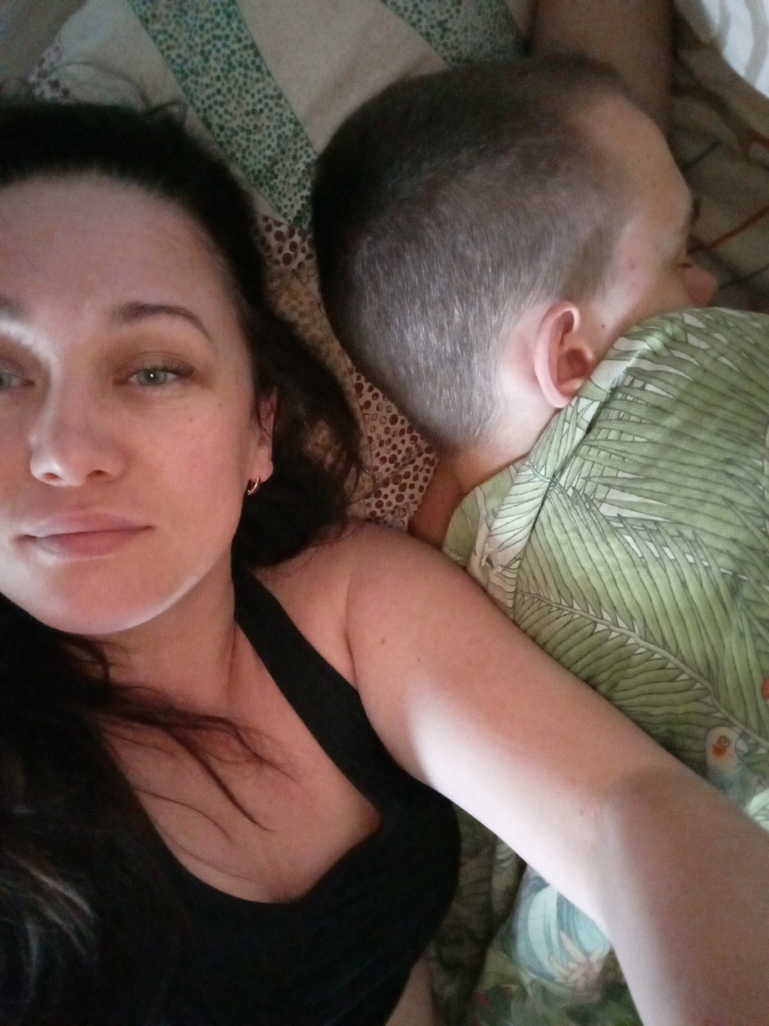 A woman with long, dark hair and light eyes is lying on a patterned bed, taking a selfie. She is wearing a black tank top. Next to her, a man with short hair is lying face down, wearing a green shirt with a tropical pattern. The location is not discernible as Chaikovsky, Russia. The environment is a bedroom.