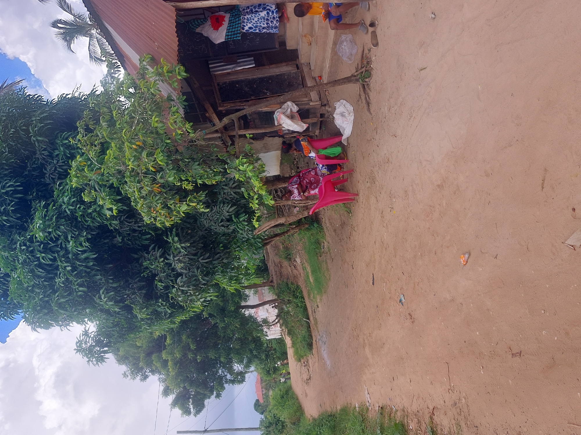 This is an outdoor scene in Dar es-Salaam, Tanzania, featuring a dirt road next to a small building with a corrugated metal roof. A lush green tree dominates the left side of the frame. Several plastic chairs are arranged near the building, where a few people are sitting. On the porch of the building, clothes hang above a blackboard, and a child sits on the steps. Trash litters the ground, and the sky is visible with scattered clouds.