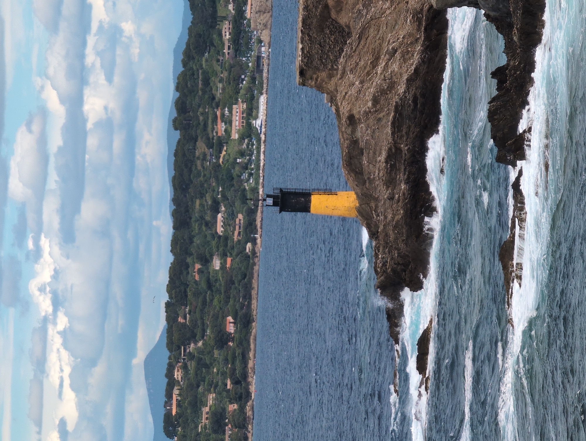 The image captures a vertical perspective of the Baie de l' Almanarre in Hyères, France. The visual composition includes a lighthouse extending from a rocky cliff, meeting the sea. The sea is rough, with white waves crashing against the rocks. On the opposite shore, a cityscape with buildings and lush vegetation is present under a partly cloudy sky.