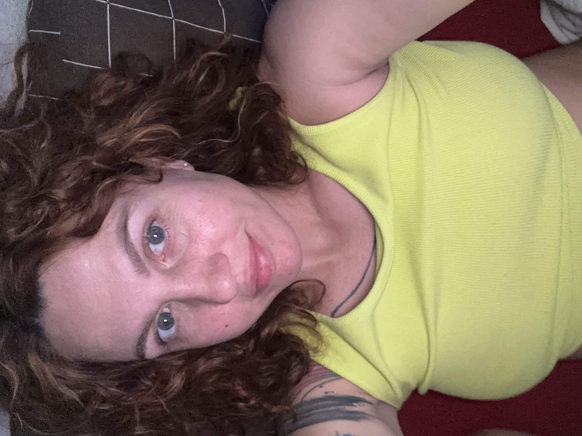 A woman with curly brown hair and blue eyes lies on what appears to be a bed. She is wearing a yellow top. A dark-colored pillow with a light-colored grid pattern is visible behind her head. A red surface, potentially a blanket or sheet, is seen under her torso. A tattoo is visible on her arm. The location is unknown and cannot be determined based on the image. It is not possible to know if this image was taken in Sochi, Russia.