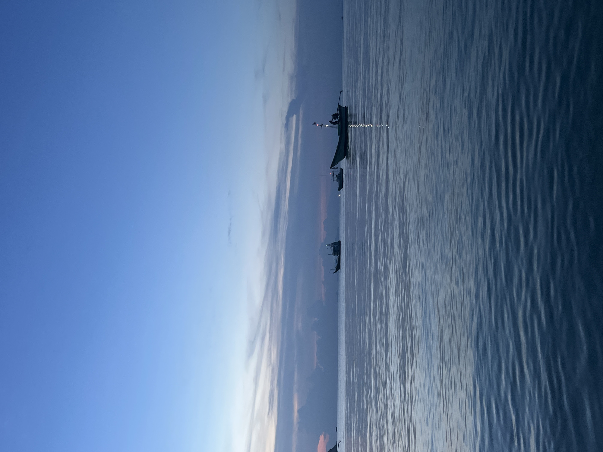 A wide shot captures the tranquil sea and sky at dusk, featuring several boats. The sky is a gradient of deep blue at the top, transitioning to lighter blues and subtle pink and orange hues near the horizon, indicating the last light of sunset or the first light of dawn. Wispy clouds are present in the lower half of the sky, adding texture. The sea is calm, with gentle ripples reflecting the dim light. In the middle ground, three boats are visible. The largest boat, on the right, is a fishing vessel with a visible light and a flag. Two smaller boats are positioned to the left of it, also appearing to be fishing boats. The overall scene conveys a sense of peace and the quiet beauty of twilight on the water. The location is Unknown, Unknown.
