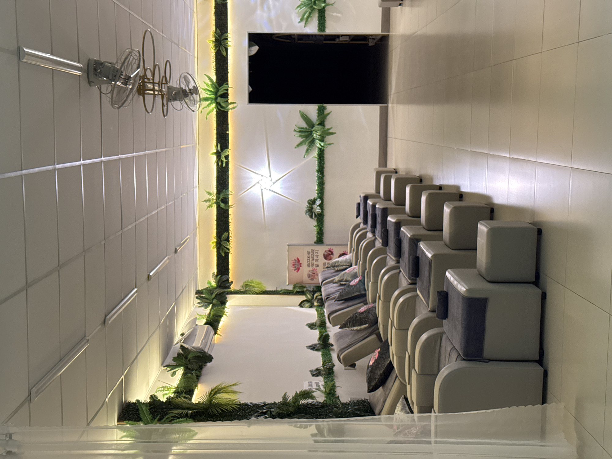 The image shows a spacious room, likely a foot massage parlor, in Nha Trang, Vietnam. The room is decorated with artificial plants along the walls and ceiling, creating a natural, spa-like atmosphere. Multiple rows of recliner chairs, upholstered in light beige, are arranged facing the wall, each equipped with a footrest. Dark gray towels are placed on the footrests and on some of the chairs. The floor is tiled with large, light-colored square tiles. In the background, a dark doorway leads to another room. Above the chairs, ceiling lights illuminate the space, and several industrial-style ceiling fans are visible. On the wall, there is a framed sign with text in multiple languages, including English, indicating "FOOT MASSAGE". The scene is quiet and unoccupied, suggesting the establishment is either closed or waiting for clients.