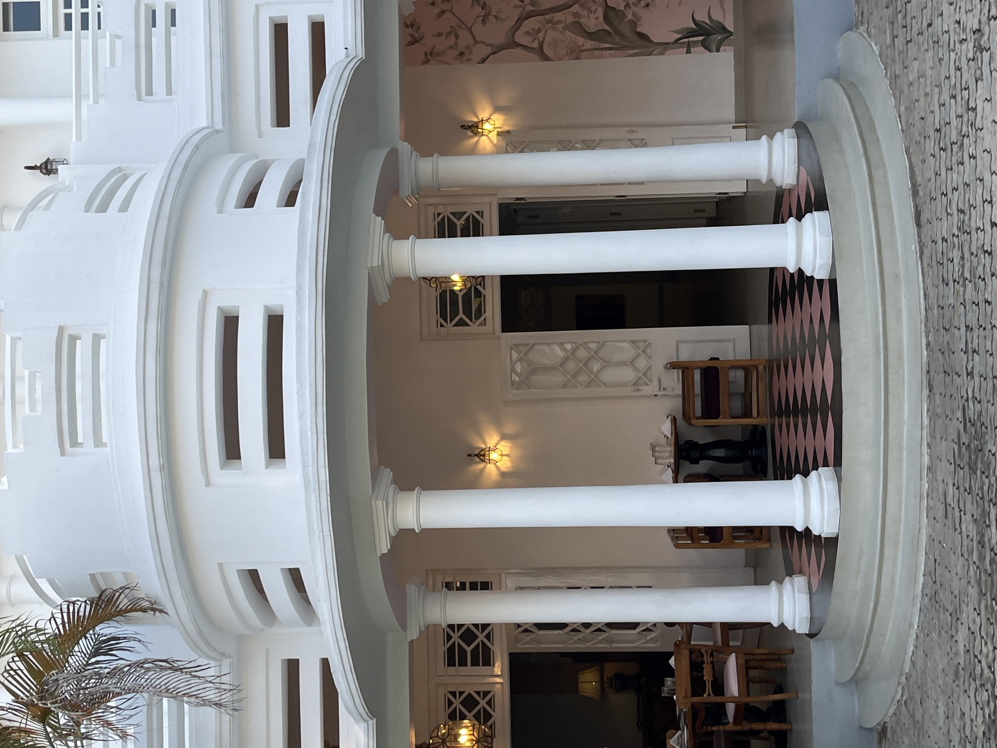 The image shows the exterior of a colonial-style building with white stucco walls and a curved balcony. There are several white pillars supporting the balcony. The entrance to the building features a patterned tiled floor in shades of pink and black. To the left, a dining table with chairs is visible through an open doorway. To the right, a wall is decorated with a floral mural. The building is located in Galle, Sri Lanka, a city known for its well-preserved colonial architecture. The lighting suggests it is daytime, with a warm ambient glow.