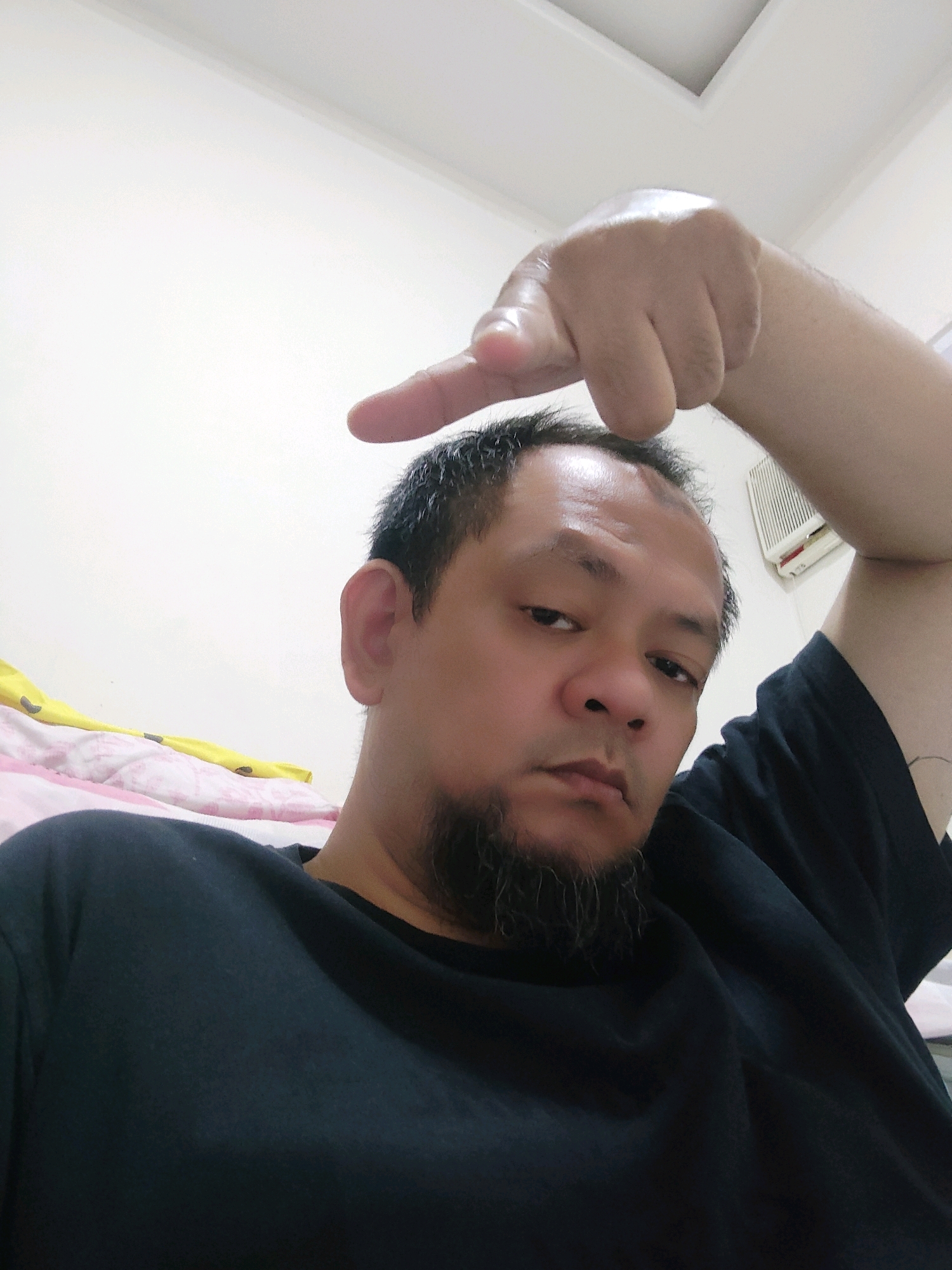 A man with short, dark hair and a goatee is taking a selfie indoors. He is wearing a black t-shirt and has his right arm raised, with his index finger pointing upwards. His left side is towards the camera, and he is looking directly into it with a neutral expression. Behind him, a white wall with a ceiling light fixture is visible, as is a portion of a bed with pink and yellow bedding. A small, white air conditioner unit is mounted on the wall in the background. The lighting suggests the photo was taken during the daytime. There is no indication of the specific location in Bogor, Indonesia.