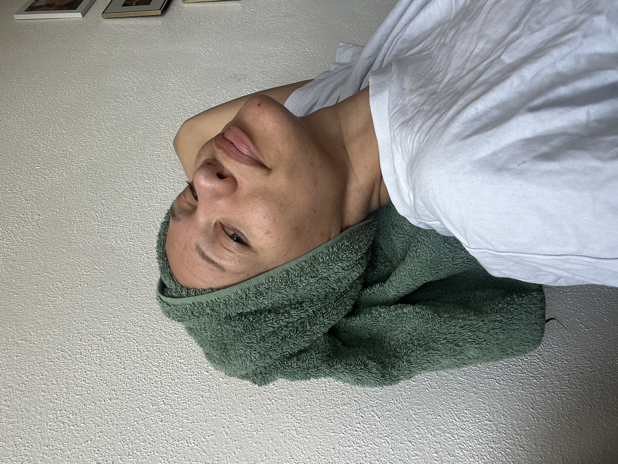 A woman wearing a light blue t-shirt and a green towel wrapped around her head is captured in a close-up portrait. She is leaning back with her arm behind her head, looking slightly upwards. The background is a textured white wall, with three framed pictures hanging on the right side of the image. The scene is indoors, suggesting a home environment. The lighting appears to be natural, coming from the right side of the frame, creating subtle shadows on her face and the wall. The overall mood is relaxed and casual. There is no information available about the specific location in Mühldorf a. Inn, Germany.