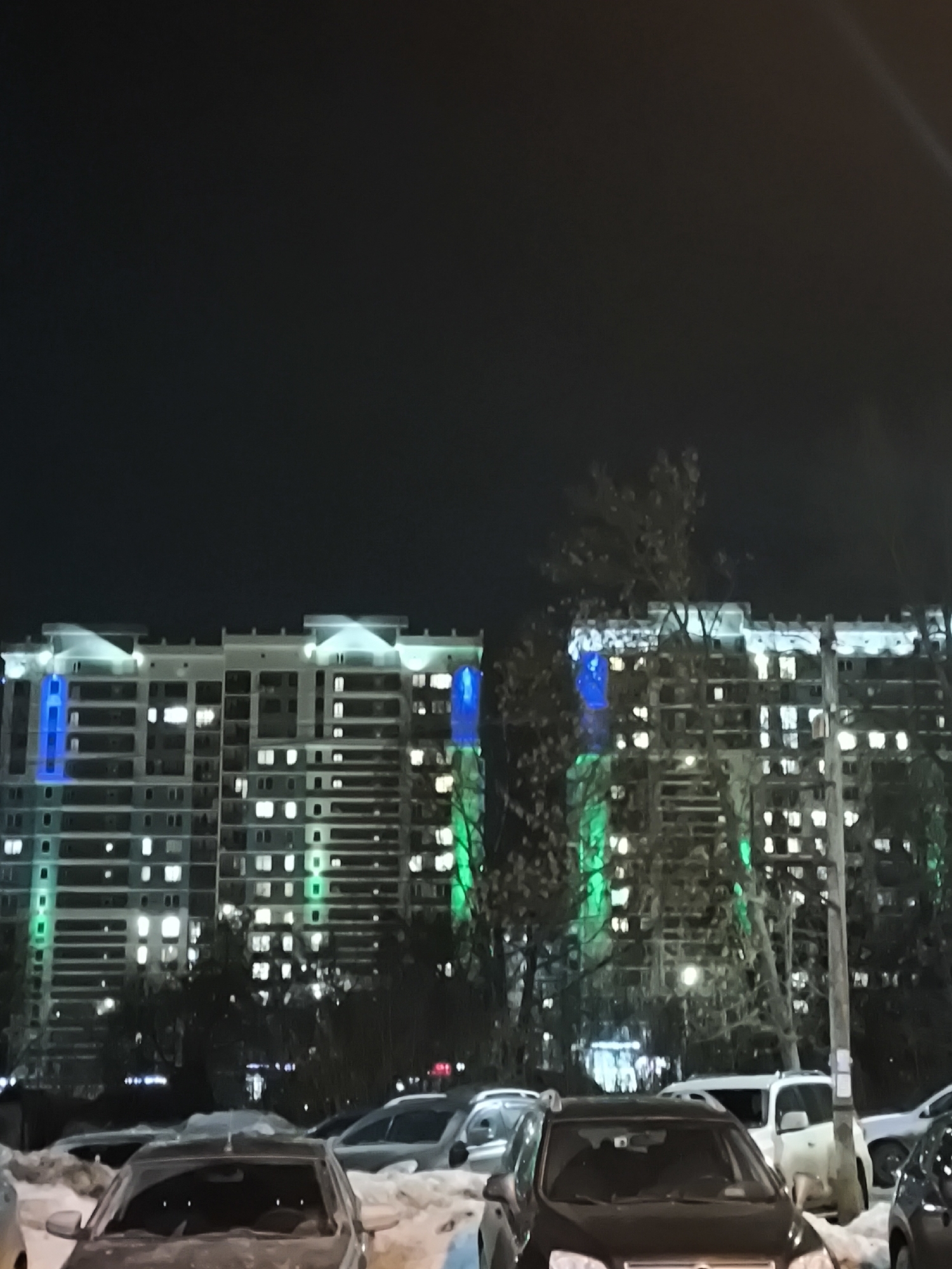 The scene depicts a nighttime exterior in Izhevsk, Russia, under winter conditions. Two multi-story residential buildings dominate the background, displaying numerous illuminated windows. The building facades are adorned with decorative lighting, including white or light blue illumination along the rooftops and distinct vertical strips of bright blue and green light on various sections of the structures. The foreground and midground feature a parking area containing multiple passenger vehicles, including sedans and SUVs, partially obscured by snow accumulated on the ground. Deciduous trees with bare branches are visible between the buildings and in the midground. No individuals are present, and no specific actions or interactions are observed.