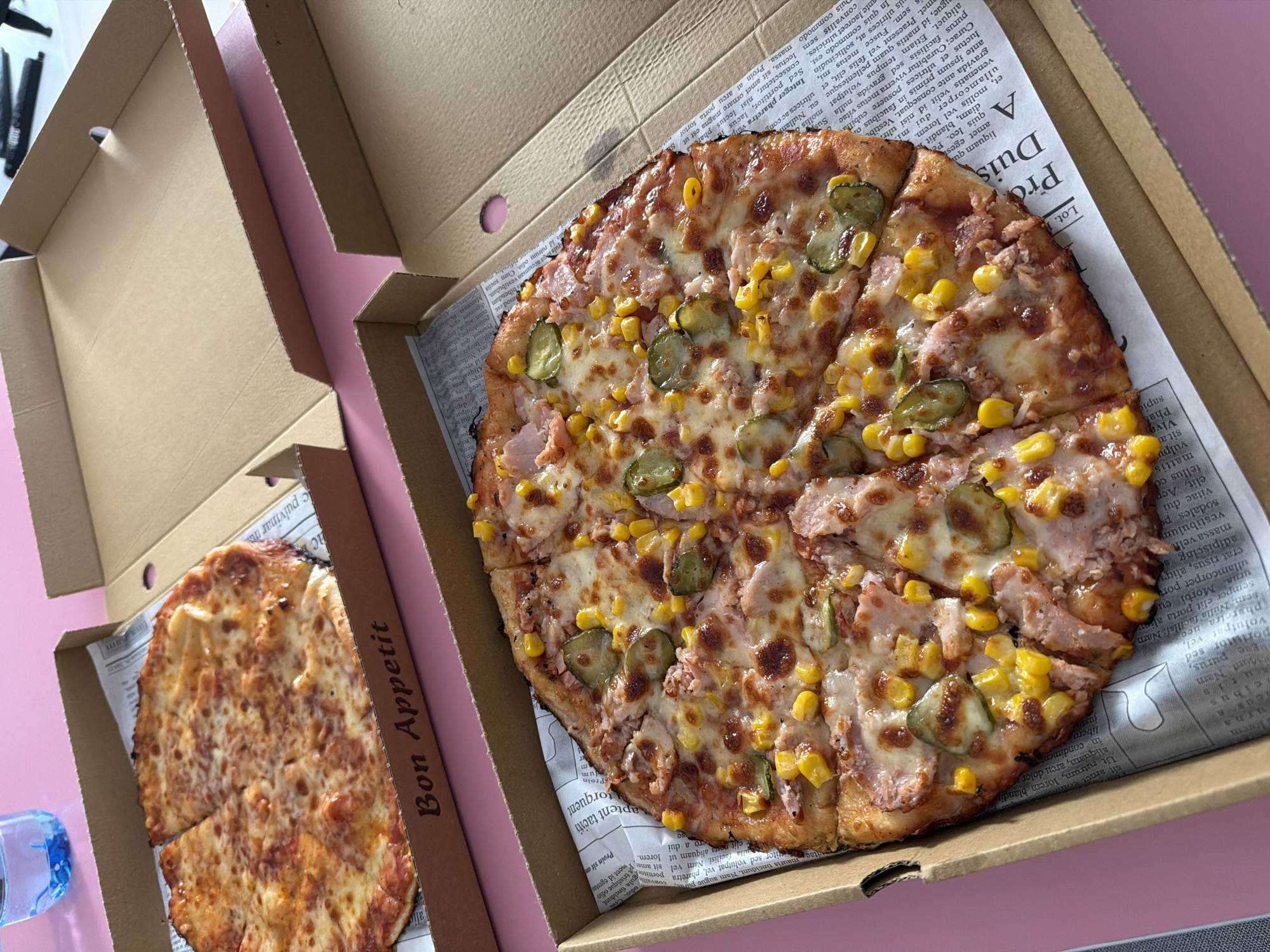 This image captures two pizzas in their cardboard boxes, set on a pink surface, suggesting a casual meal or takeout experience, likely indoors during daylight.
The dominant subject is a large, round pizza on the right, cut into multiple slices. It features a generous topping of melted cheese, bright yellow corn kernels, distinct green pickle slices, and what appears to be shredded ham or another meat. This pizza rests on a piece of paper resembling a newspaper within its brown cardboard box. The visible text on this paper is largely Latin "Lorem Ipsum" placeholder text, including prominent headings like "A Duis" and "Pro," indicating it's a generic design rather than a real newspaper.
To the left, a smaller, round pizza, also sliced, is partially visible in its own brown cardboard box. This pizza appears to be a classic cheese variety, with melted cheese and hints of sauce. The side flap of this box clearly displays "Bon Appetit."
A transparent plastic water bottle is partially visible in the bottom-left corner of the frame. The overall scene conveys a relaxed atmosphere around a ready-to-eat meal. The image does not contain specific visual or textual cues to confirm the location as Sofia, Bulgaria.