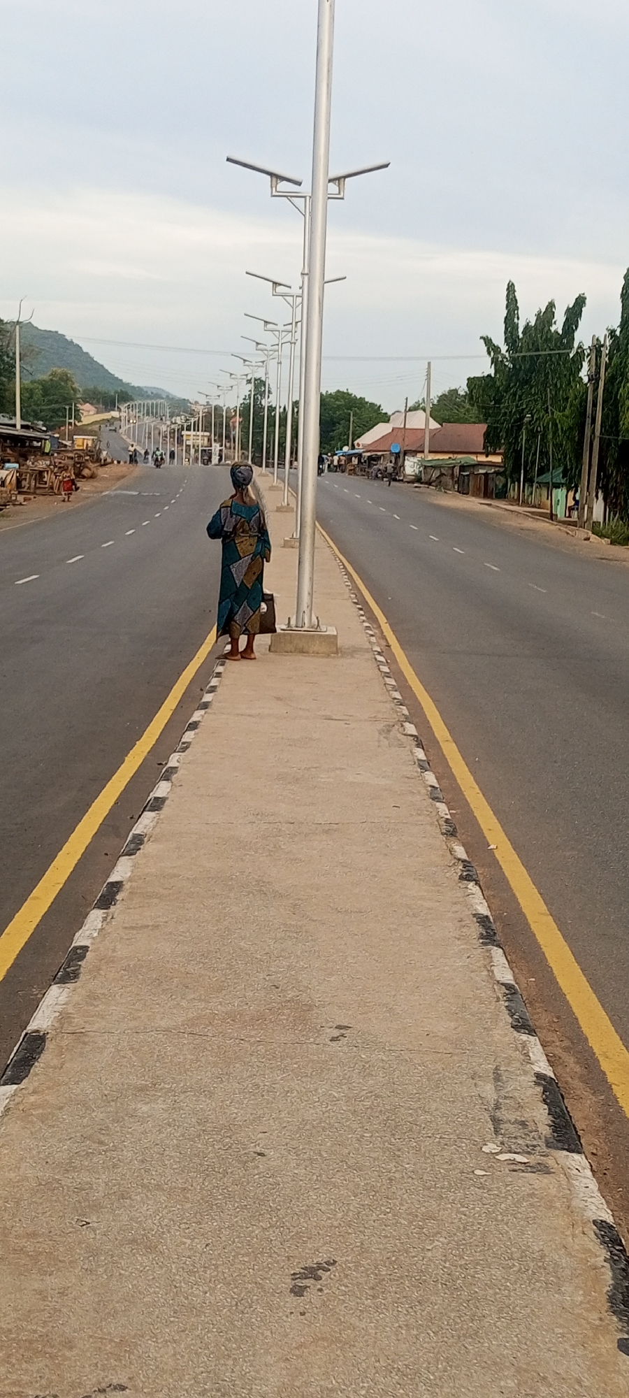 An image of a woman standing on a median strip of a highway in Zango-ndaji, Nigeria. The woman faces away from the camera, dressed in a blue and brown patterned garment and carrying a brown bag. A series of silver streetlights line the median strip. The highway is flanked by yellow lines and features intermittent white and black markings on the median’s edge. Buildings and trees are visible on either side of the road. Distant hills can be seen in the background under a cloudy sky.