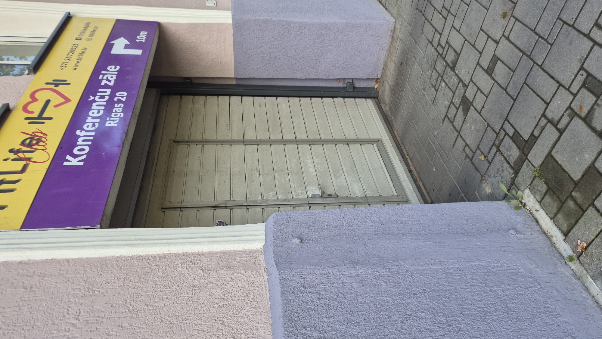 The image shows the entrance to a business labeled "FitLife Club" in Daugavpils, Latvia. The business name is displayed on a yellow and purple sign above a closed, light-colored, slatted roll-up door. Below the "FitLife Club" logo, the purple section of the sign reads "Konferenču zāle" (conference hall) and "Rīgas 20" (Riga Street 20). To the right of this text, a white arrow points to the right, with "10m" written below it, indicating a distance. The entrance is flanked by building walls, one painted in a textured light pink and the other in a textured lavender. The ground in front of the entrance is paved with gray interlocking bricks. A small patch of greenery is visible growing from the ground near the left corner of the building. The scene appears to be an exterior shot of a commercial establishment on a street.