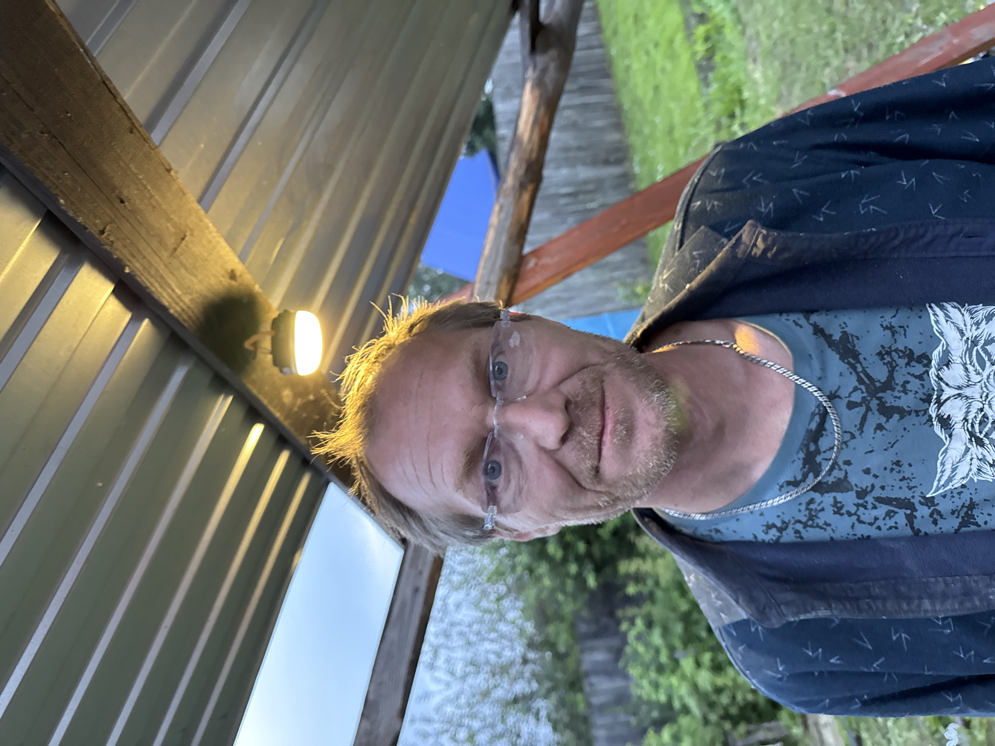 An outdoor medium shot captures a man in his late 40s or early 50s, with light brown hair, blue eyes, and a light beard, wearing glasses. He is positioned under a metal roof supported by wooden beams, with a light fixture hanging from one beam. The background shows a wooden fence, green grass, and trees, suggesting a suburban or rural setting. The man is wearing a dark blue patterned jacket over a blue t-shirt with a graphic print and a silver chain necklace. The lighting is warm, likely from the overhead fixture and the setting sun. The scene appears to be in Chaikovsky, Russia, given the provided context. There is no discernable action or interaction taking place.