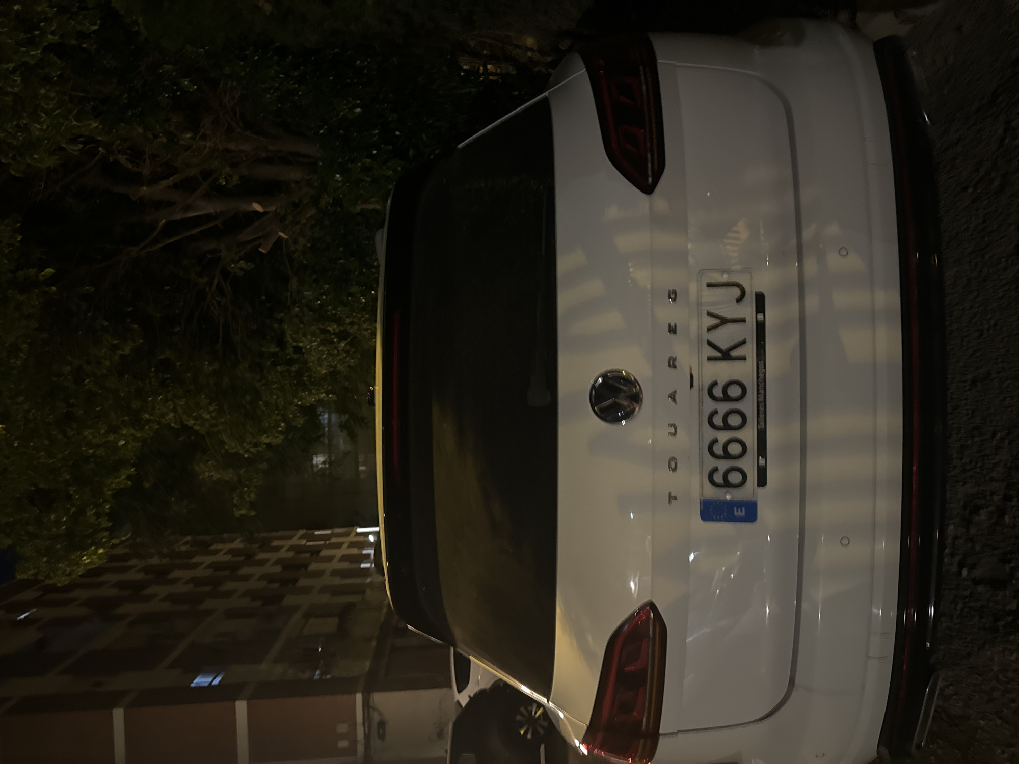 A white Volkswagen Touareg is parked on a street at night. The car's rear is facing the camera. The license plate reads "6666 KYJ". A building with multiple floors and windows is visible on the left side of the image, and trees with dark foliage are on the right. The scene appears to be in an urban environment.