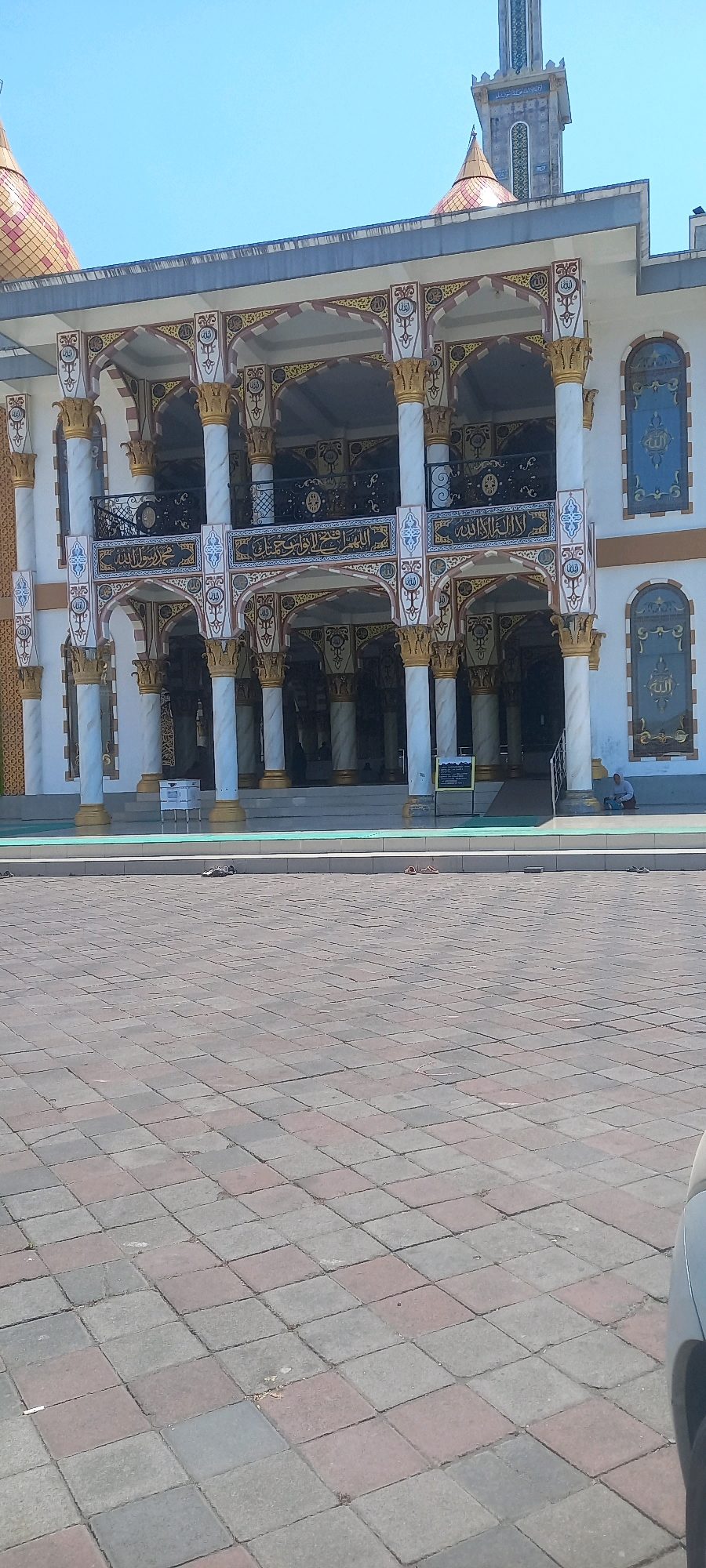 The image shows the facade of the Masjid Al-Mutaqin, located on Jl. Kalimantan, Jember, Indonesia. The mosque features a white exterior with ornate gold and maroon detailing on the arches and pillars. Two golden domes are visible on the roof. In front of the mosque is a paved courtyard made of square interlocking bricks in various shades of gray, red, and beige. A person wearing a light-colored headscarf is seated on the ground to the right of the mosque entrance, appearing to be engaged in prayer or contemplation. In the foreground, several pairs of sandals are scattered on the paving stones, indicating recent use by mosque visitors. The sky above is clear and blue.