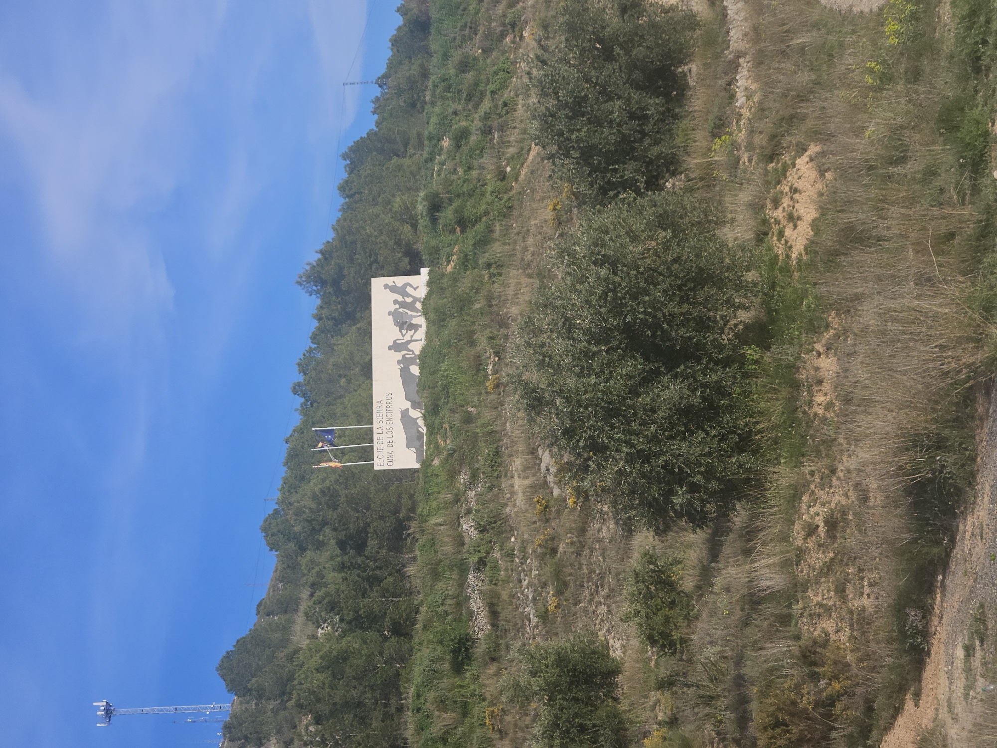 The image shows a hillside in Elche de la Sierra, Spain. A white monument stands prominently on the hillside, depicting a silhouette of figures running. Text on the monument reads "ELCHE DE LA SIERRA, CUNA DE LOS ENCIERROS". Two flags fly next to the monument. The hillside is covered with trees, brush, and some exposed soil. A tall communication tower is visible in the lower left corner of the image. The sky is blue. A road or path is visible on the right side.