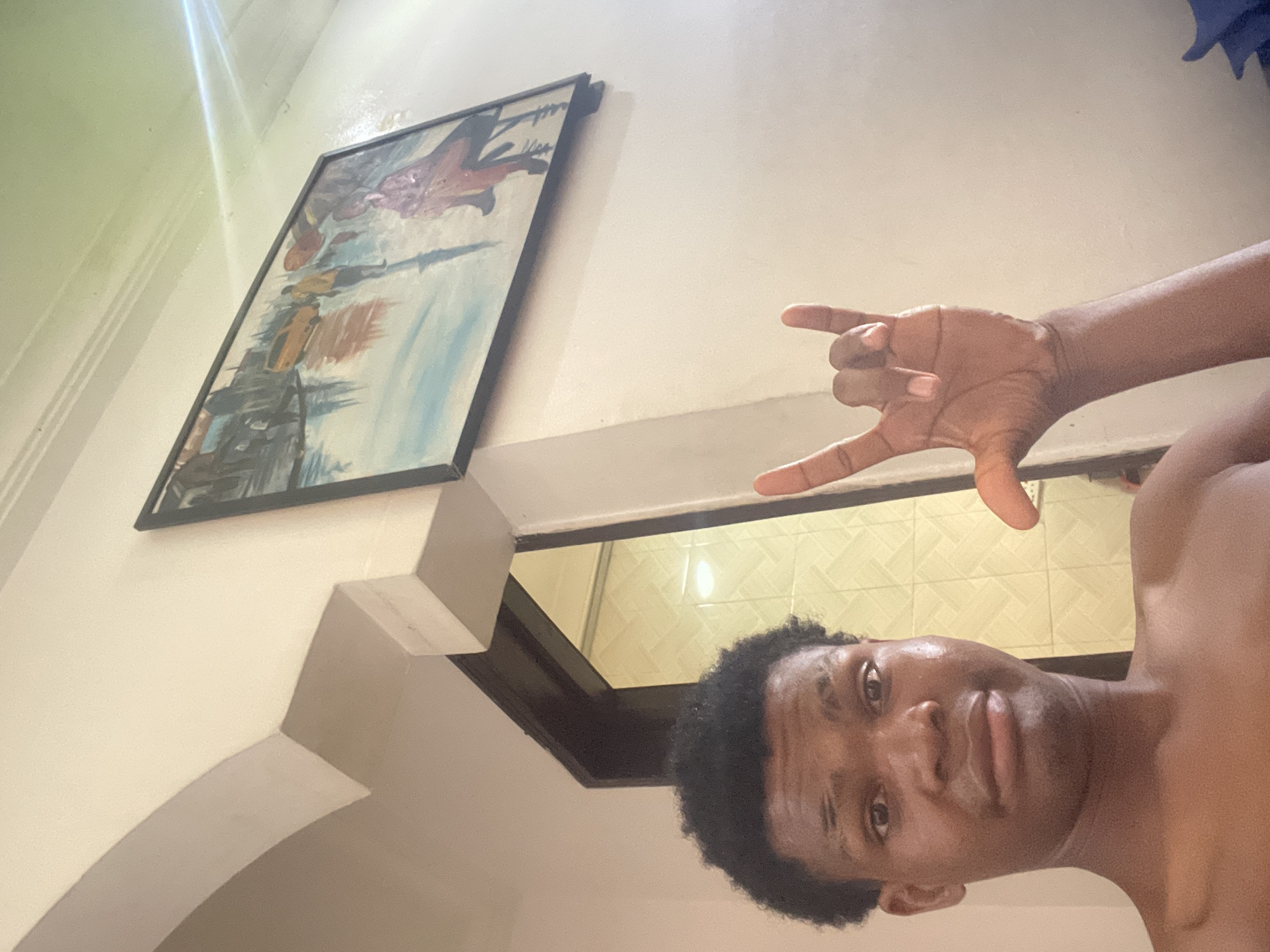 A young Black male with short, curly hair is taking a selfie in what appears to be a residential setting in Ado, Nigeria. He is shirtless, and his left hand is raised, forming a rock-and-roll sign with his index and pinky fingers extended. His facial expression is a slight grimace. Behind him, to the right, a framed painting depicting a street scene with a yellow taxi and figures walking in what appears to be wet conditions hangs on a lightly textured, cream-colored wall. To the left, an arched doorway leads into a room with tiled walls. The lighting suggests it is daytime, with a strong light source creating a glare in the upper right portion of the image. The overall environment seems casual and domestic.