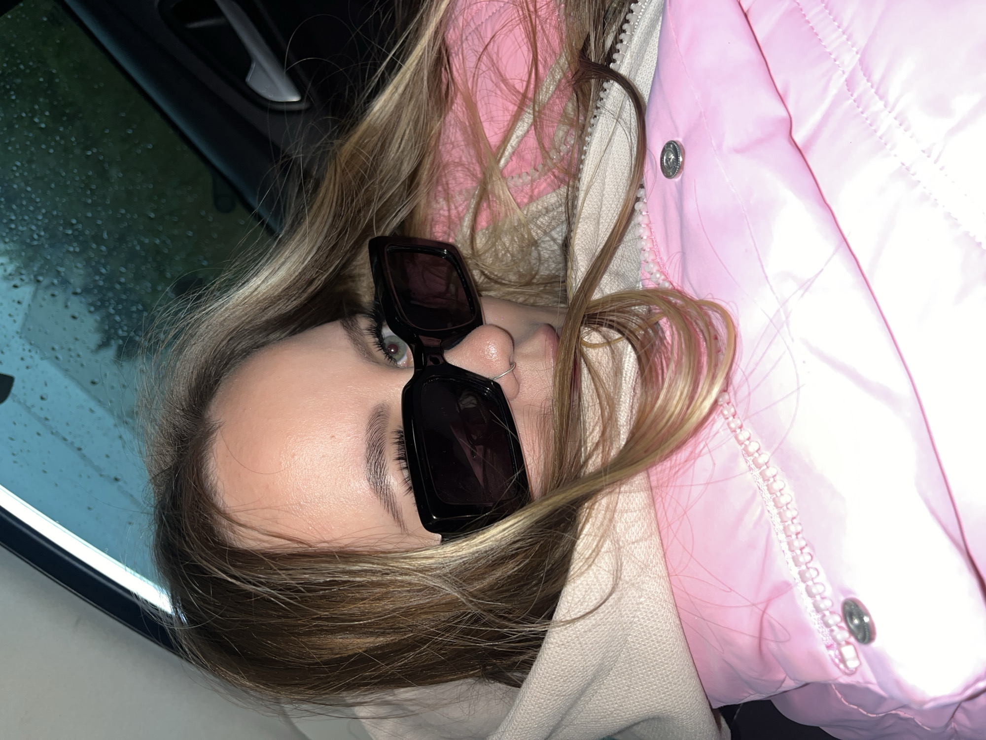 An upward-angled, close-up shot captures a person, likely a young woman, lying down or leaning back inside a vehicle. Her head is tilted, and her long, light brown hair, with blonde highlights, frames her face and spills around her. She wears large, dark, rectangular sunglasses that obscure her eyes, and a small nose ring is visible.
She is dressed in a bright pink, quilted puffer jacket with a visible zipper and a silver snap button, worn over a light-colored, possibly hooded, garment. To the left, a car window is visible, covered in numerous small water droplets, indicating rain or recent rain. The background seen through the window appears dark, suggesting it is nighttime or very low light conditions outside. A car door handle is partially visible in the upper part of the frame.
The overall impression is a casual, relaxed pose, possibly a selfie, taken in a car during or after rainfall. There is no visible text in the image. The provided location context for this image is Goryachiy Klyuch, Russia.