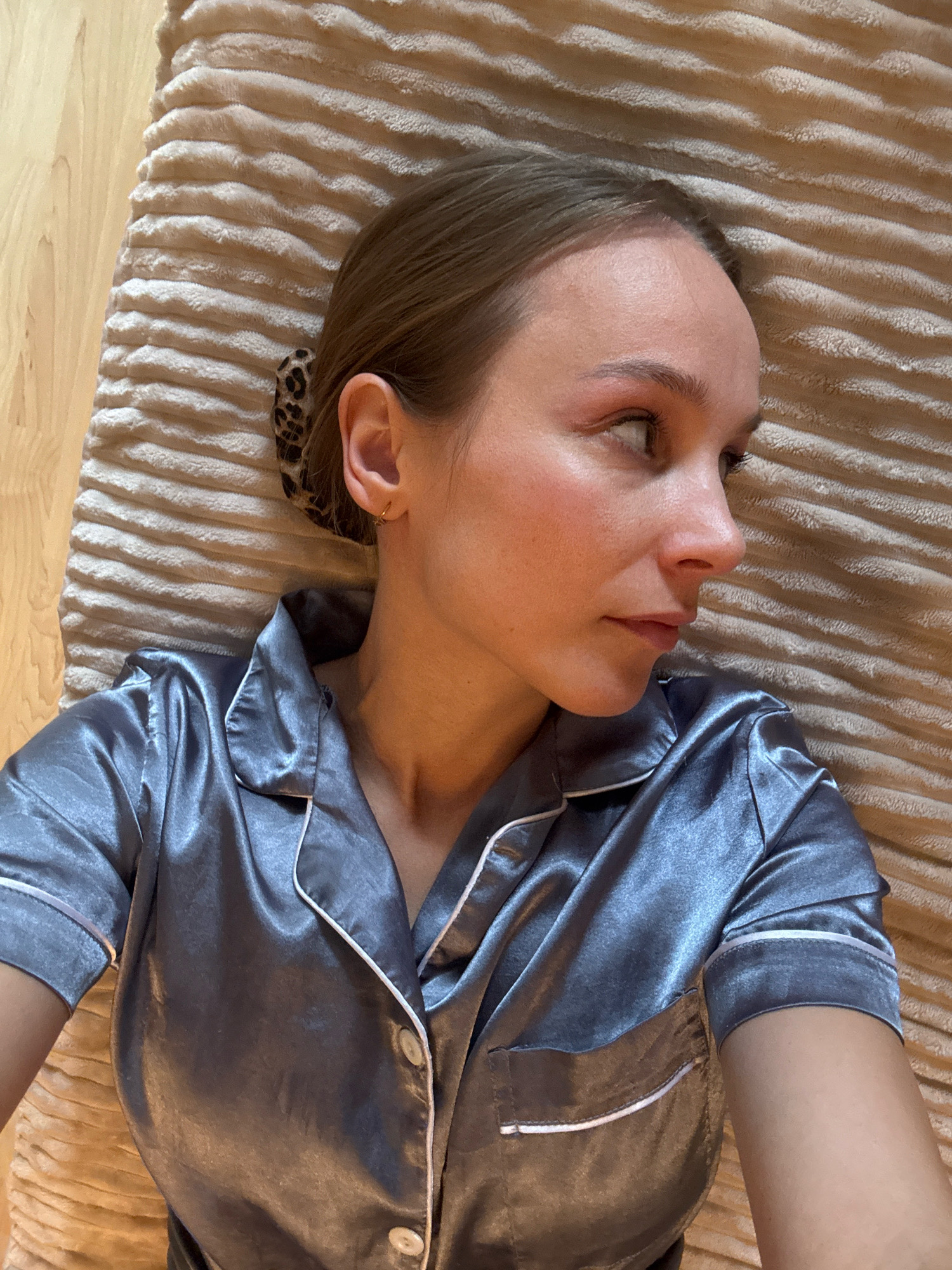 A young Caucasian woman with light brown hair styled into a bun with a leopard print hair accessory, and a small gold hoop earring, lies on her side. She is wearing a gray satin pajama top with white piping and two pearl buttons. The woman is looking to her right, with her head resting on a textured, beige blanket. The background shows a wooden floor. The image appears to have been taken indoors, likely in a bedroom or living space in Überherrn, Germany.