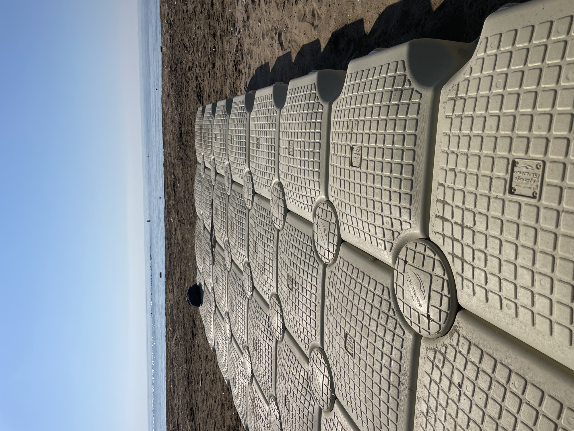 In Trikomo, Northern Cyprus, a series of interlocking modular plastic floats forms a pathway on a sandy beach leading towards the sea. The floats are a light tan color and have a textured grid pattern on their surface. A dark blue baseball cap rests on the floats in the foreground. Beyond the floats, the sandy beach transitions to the calm, blue sea under a clear sky. The horizon is visible with a faint line of land in the distance.