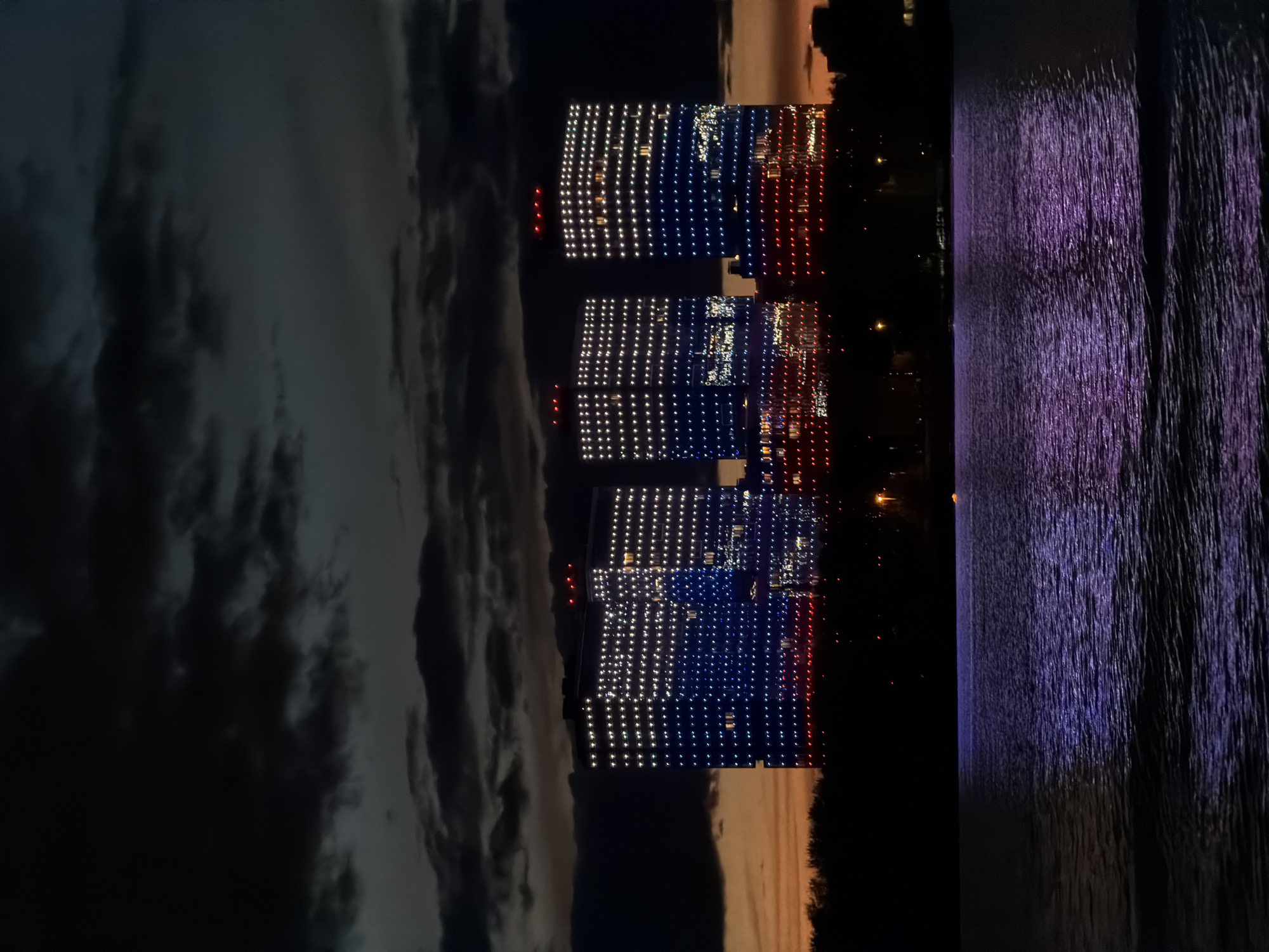 At night, three illuminated buildings in Kazan, Russia, display a color pattern of white, blue, and red. To the left, dark clouds hang above a twilight-lit horizon. To the right, purple light reflects on water. Buildings and streetlights provide additional light.