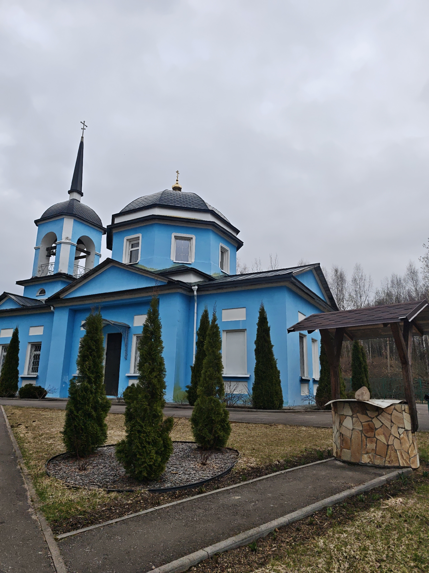 The image depicts a bright blue building, likely a church, with two domes topped with crosses, located in Nadezhdino, Russia. A black steeple-like structure with a cross atop stands next to a larger, more rounded dome. The building has white window frames and architectural accents. In front of the building are several tall, slender evergreen trees planted in a mulched bed, and a paved path leads to a stone well structure with a wooden roof. The sky is overcast and gray, suggesting a cloudy day. The surrounding area appears to be a lawn or grassy area, and bare trees are visible in the background, indicating a fall or winter season. The image is taken from a ground-level perspective. No people are visible.
