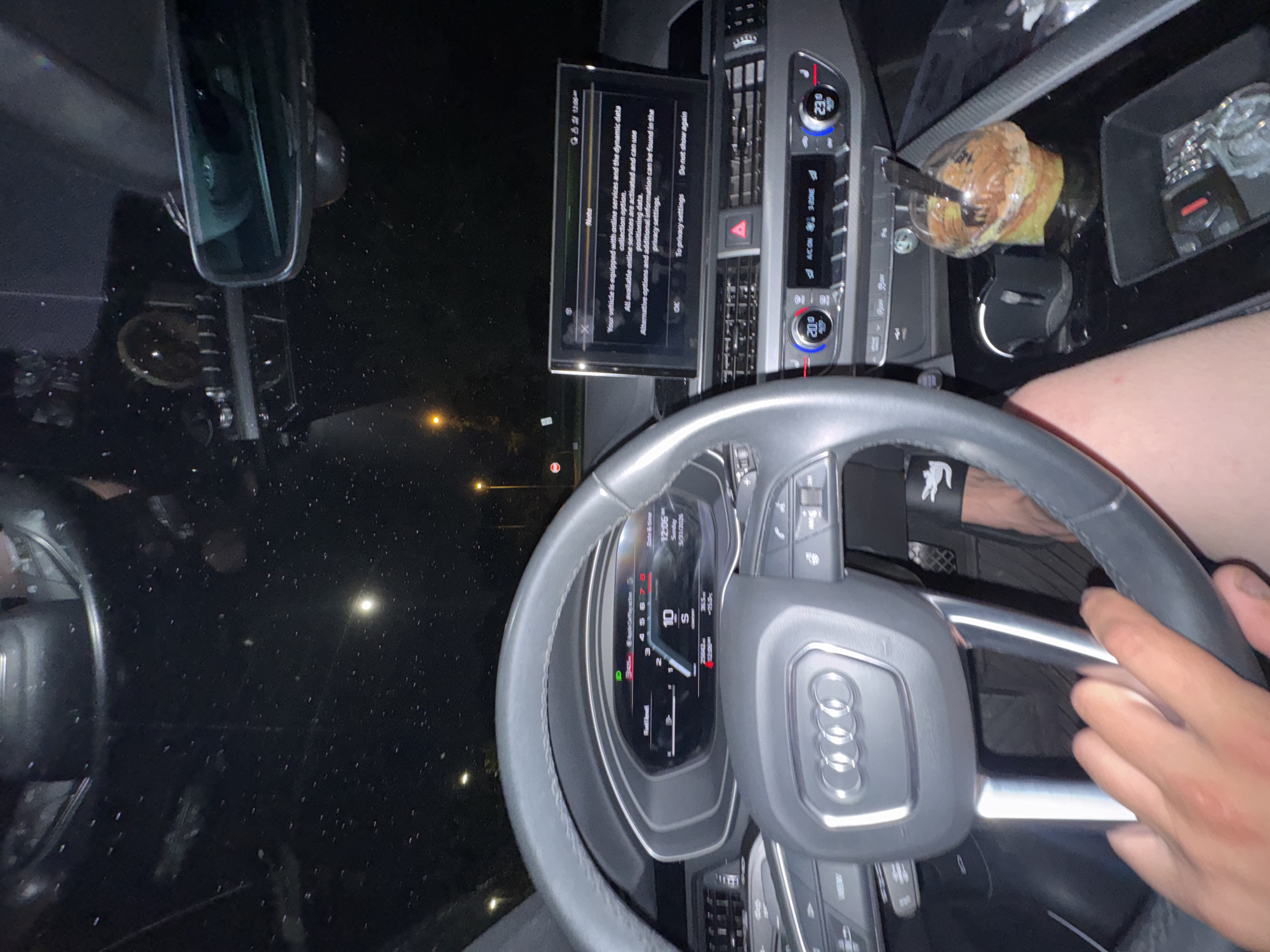 The image shows the interior of a parked Audi car at night. The steering wheel is in the foreground, with the Audi logo visible. The dashboard features a digital display showing a notification about online services and data collection. The car's infotainment system displays climate control settings. A cup with a straw sits in the cup holder. The driver is holding the steering wheel. The location cannot be determined from the image. The location of Schaumburg, United States of America is irrelevant to the context of the image.