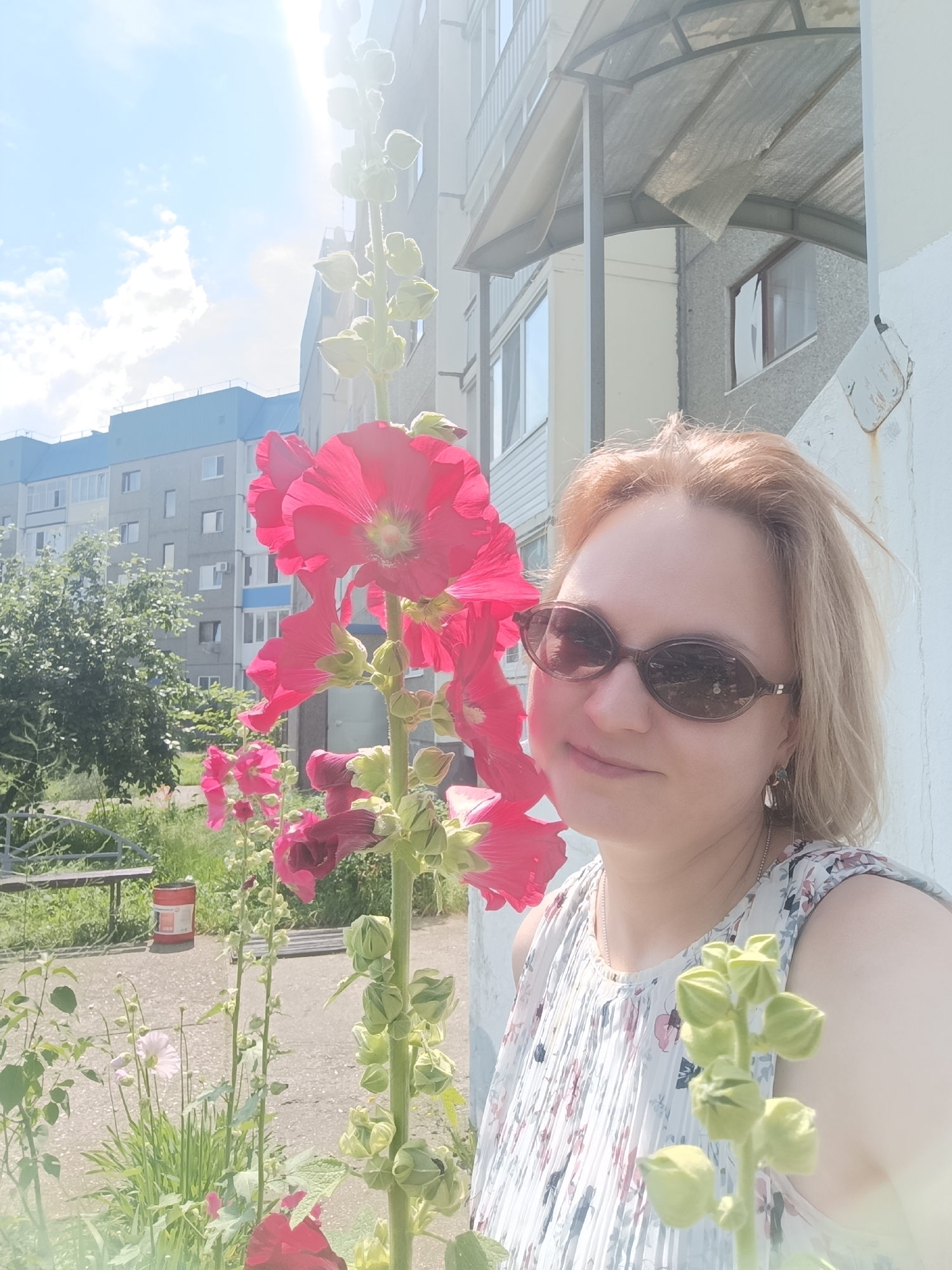 A woman with blonde hair and sunglasses is taking a selfie in front of a building in the city of Markovsky, Russia. She is wearing a floral print dress and a necklace. To her left, there are large pink flowers with green buds. In the background, there is a multi-story apartment building with blue accents and trees. The sky is blue with some clouds, and the sun is shining brightly, causing lens flare.