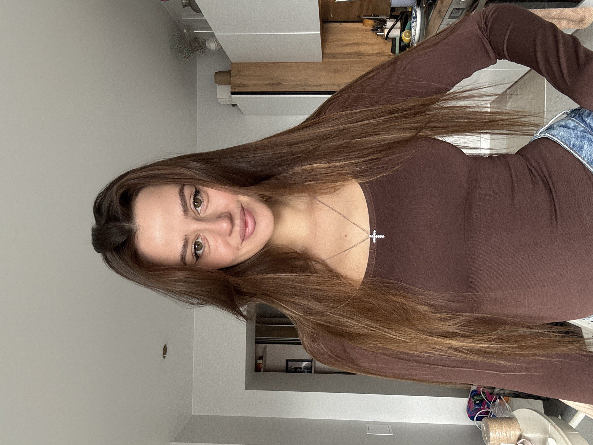 A woman with long brown hair, wearing a brown long-sleeved shirt and blue denim shorts, is looking at the camera. She is wearing a silver necklace with a cross pendant. The location appears to be an indoor space with white walls. Some kitchen items are visible in the background, including a white cabinet, a wooden counter, and some utensils. It's not possible to confirm the specific location in the city of Krasnoyarsk, Russia.