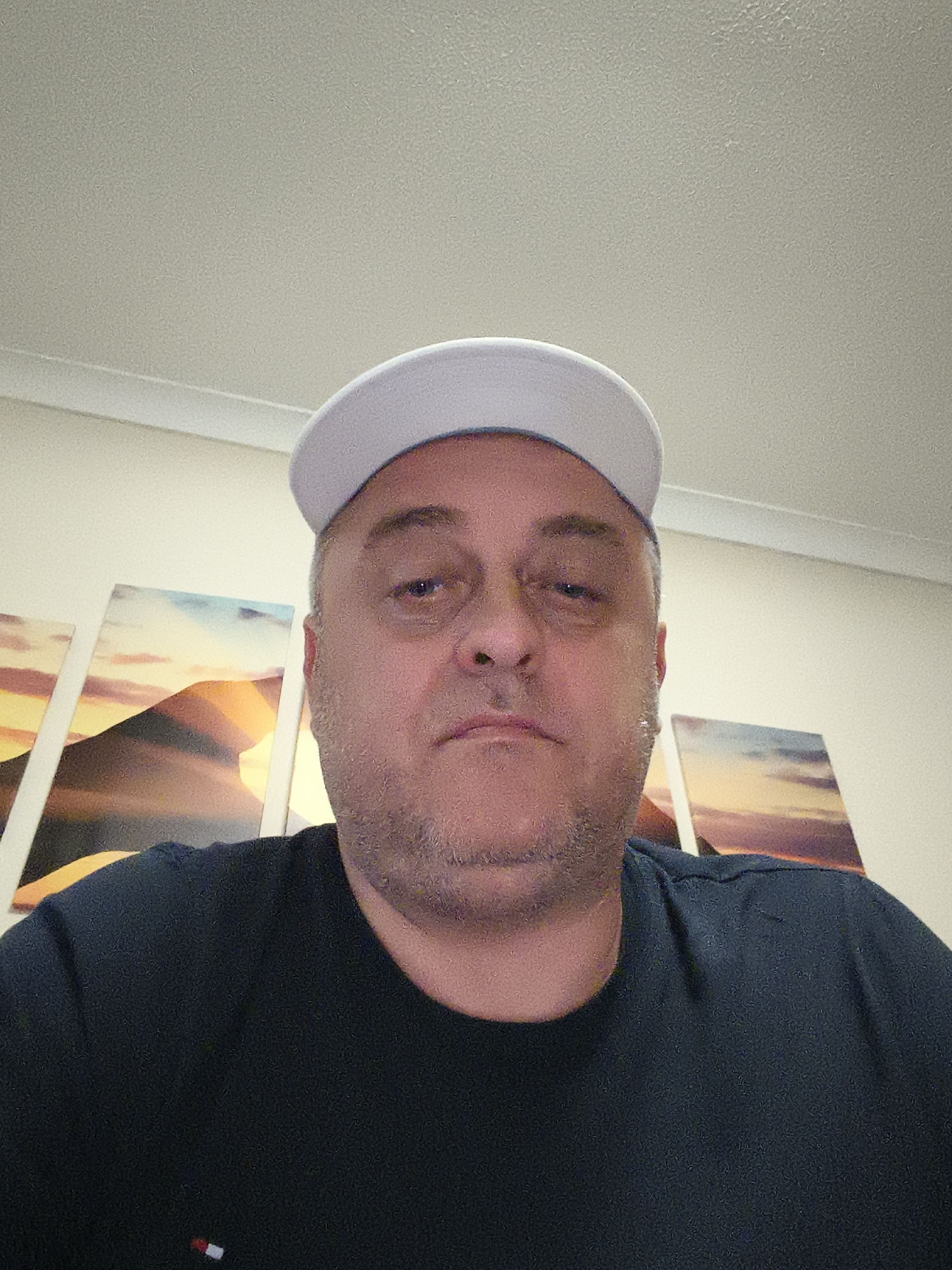 A middle-aged man is depicted in a medium close-up, head-and-shoulders portrait shot indoors. He has short, greying hair and noticeable stubble on his face. He wears a light-colored (possibly white or light grey) baseball cap and a dark blue or black crew-neck t-shirt. A small, rectangular logo with red, white, and blue sections, resembling the Tommy Hilfiger brand emblem, is visible on the left side of his chest. His expression is neutral, appearing somewhat serious or slightly tired, and he looks directly into the camera.
The background features a light-colored, possibly cream or off-white, wall. Mounted on the wall are three framed art pieces, or canvases, arranged horizontally. These artworks abstractly portray desert or mountainous landscapes, characterized by warm tones of orange, yellow, and red blending into cooler blues and purples, evocative of a sunset or sunrise. The ceiling, visible in the upper portion of the image, is also light-colored and exhibits a textured surface. The lighting suggests an interior setting with artificial illumination. The man is likely taking a selfie or being photographed, with no other activity apparent. The scene is consistent with a residential interior located in Huntingdonshire, United Kingdom.