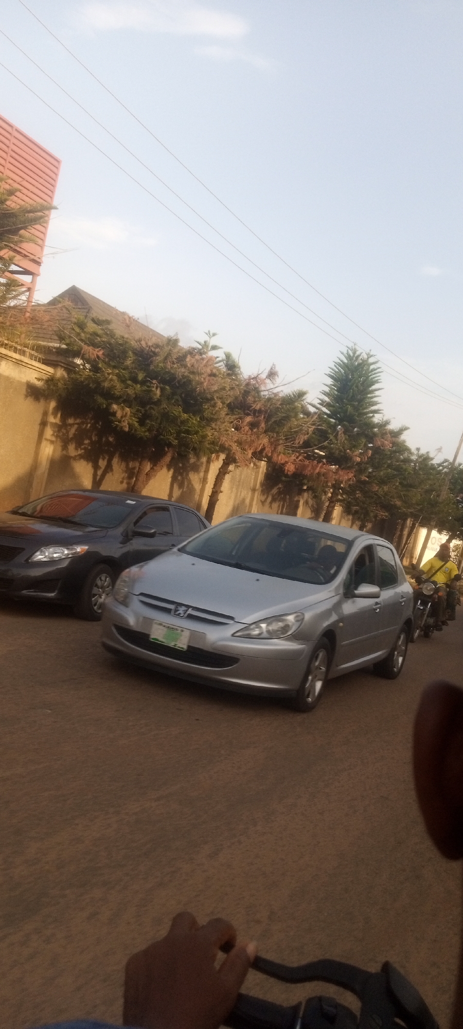 The photograph shows a street scene in Narayi, Nigeria. A silver Peugeot 307 is parked on the side of the road alongside a dark-colored Toyota Corolla. A man on a motorcycle is positioned near the Peugeot. The surroundings consist of a building, a concrete wall, and trees. Part of a hand on motorcycle handlebars appears in the lower part of the photograph, suggesting the photo was taken from a moving motorcycle. Utility lines are visible against the sky.