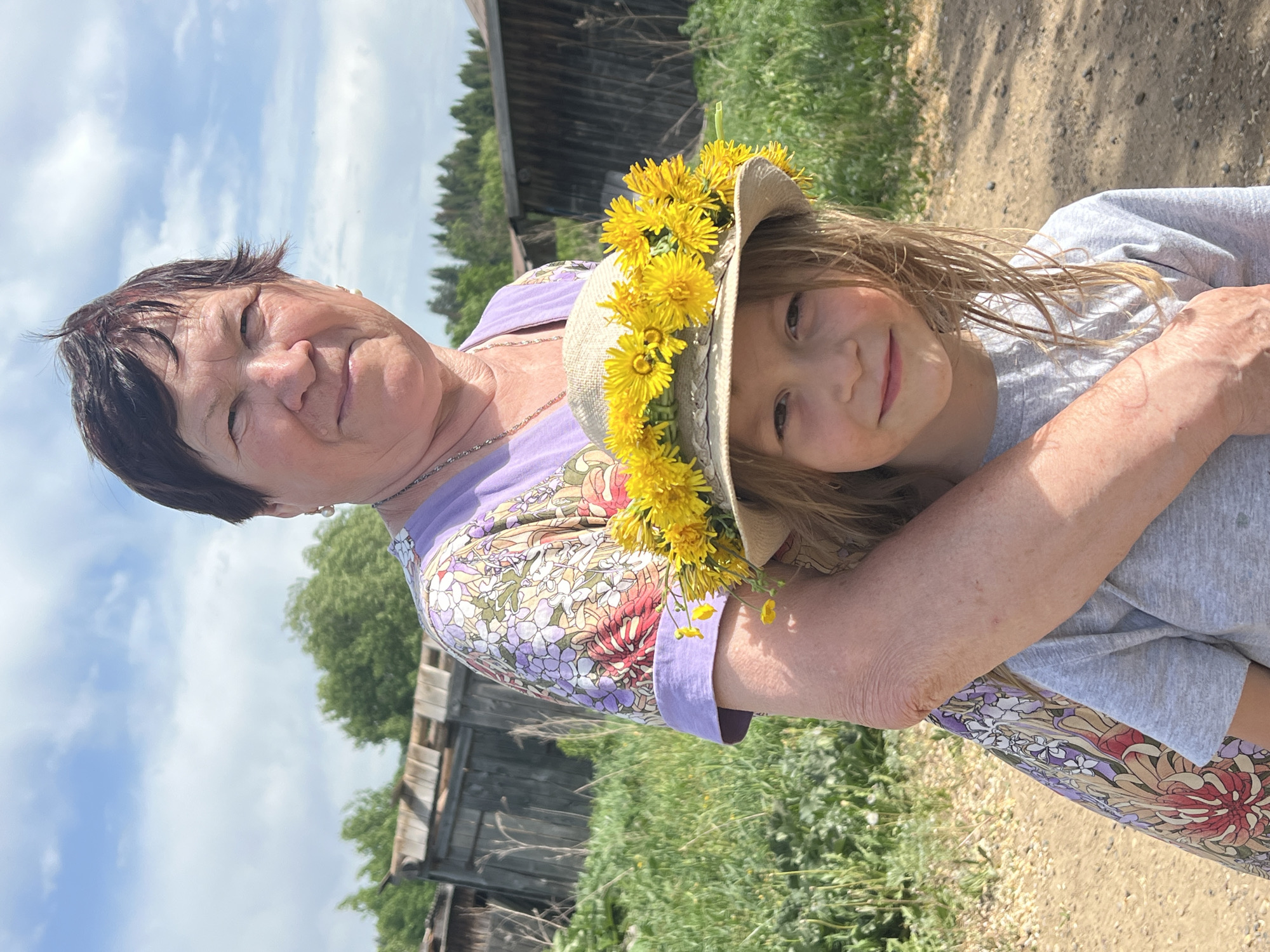 A woman and a young girl stand embracing in an outdoor setting, likely near or in Panteleevka, Russia based on the prompt. The woman, presumably older, has short dark hair and is wearing a floral-patterned shirt with a lilac trim. The young girl has light blonde hair, some of which is tucked under a straw hat adorned with yellow dandelions. The woman's arm is around the girl, who is wearing a plain grey top. Behind them is a weathered wooden structure and a patch of green vegetation. The sky is partly cloudy. The ground appears to be a dirt path.