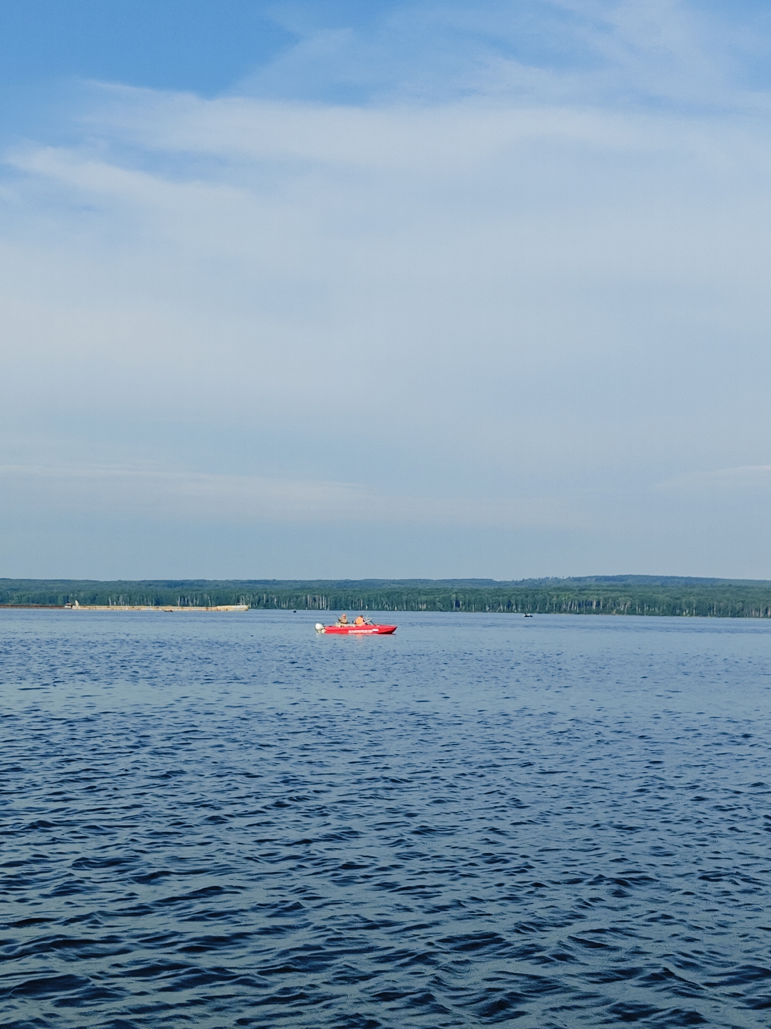 The image depicts a wide, calm body of water, likely a reservoir or a large river, under a partly cloudy sky. In the middle ground, a bright red boat with at least two people is visible. The boat appears to be a small motorboat. On the horizon, a long, flat, lighter-colored barge or floating structure is being towed by an unseen vessel, with a dense line of green trees forming the backdrop of the landscape. The water is deep blue with gentle ripples. This scene is consistent with the Kama River reservoir in the city of Chaikovsky, Russia, which is known for its industrial activity and recreational use of the water.