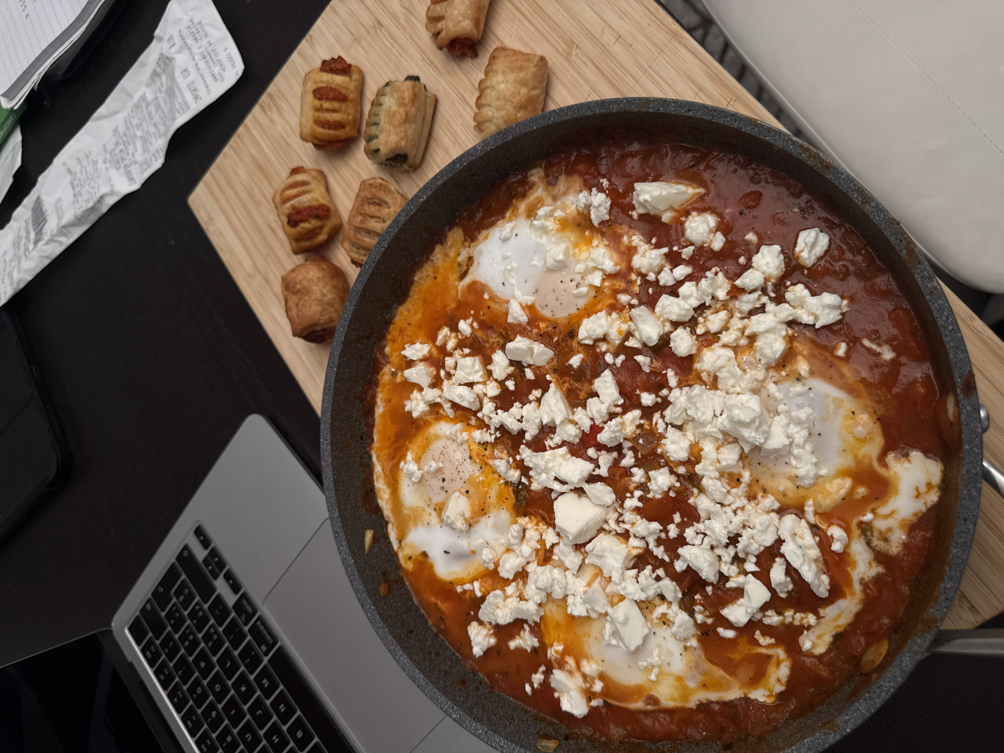 This overhead shot captures a meal laid out on a dark surface, likely a desk or table, in an indoor setting in Milan, Italy. The focal point is a dark, round pan containing a vibrant red, tomato-based dish, resembling shakshuka. Four cooked eggs are visible within the sauce, generously topped with crumbled white cheese, consistent with feta.
To the upper left of the pan, on a light wooden cutting board, are several small, golden-brown pastry rolls. Some rolls show a reddish filling, while one distinctively has a green filling, possibly spinach.
Further to the left, a crumpled white paper receipt is visible. Partially legible text on the receipt includes "PREZZI," "SERVIZIO," and the currency "EUR," indicating a transaction in Italy. Below the receipt, a silver-grey laptop, likely an Apple MacBook, is partially visible, displaying its dark keyboard. A dark smartphone or tablet is also partially obscured next to the laptop. A segment of a white chair or piece of furniture is seen in the upper right background. The scene suggests a casual meal, possibly breakfast or brunch, enjoyed in a work or home environment.