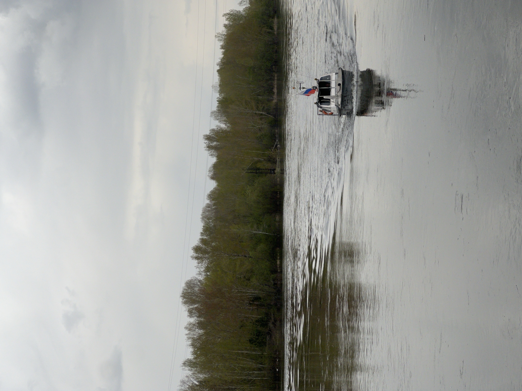 A boat is moving along the Yenisey River in Krasnoyarsk, Russia. The boat has a Russian flag. The trees lining the riverbank are mostly bare. It is an overcast day.