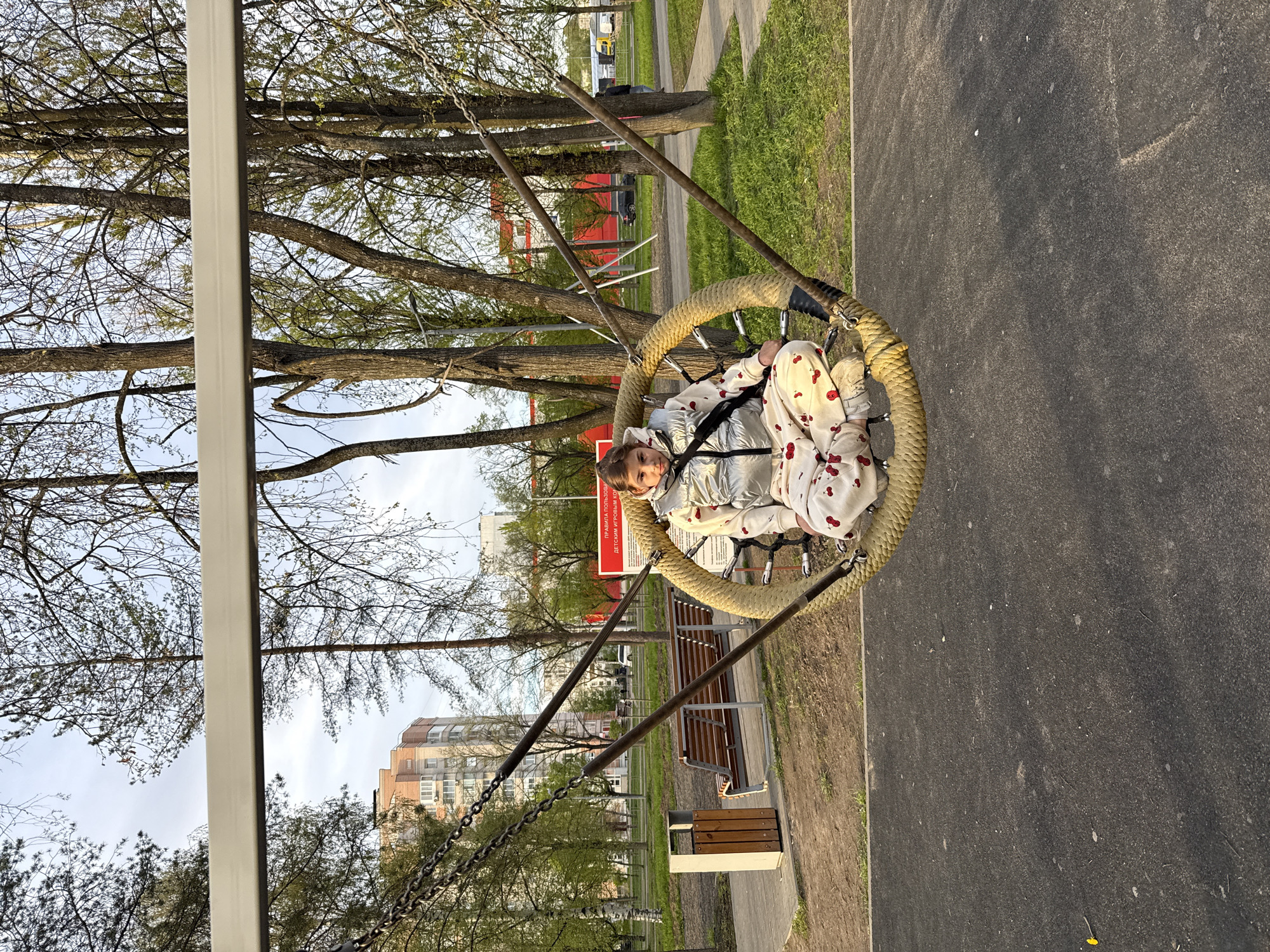 A young girl is sitting in a round swing on a playground in Rusanovo, Russia. She is wearing a silver jacket and cherry-print pants. The swing is suspended by chains attached to a metal frame. Behind the swing are trees, a red sign with white Cyrillic text, and a bench. In the background, there are apartment buildings, indicating a residential area. The playground is situated next to a paved path, and grass is visible in the surrounding area.