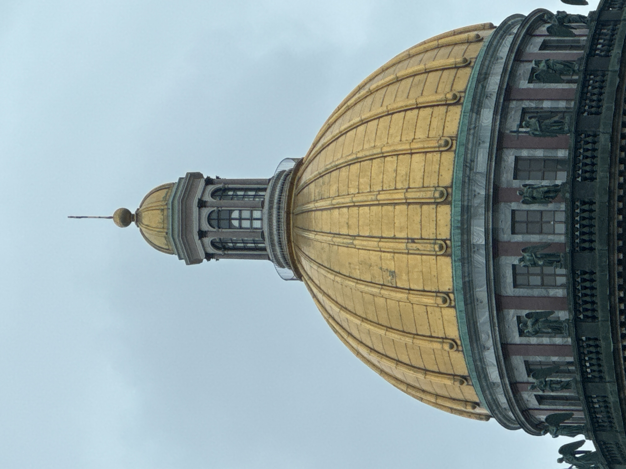 This media file presents a close-up, upward view of the gilded dome and upper structures of a grand architectural building, set against an overcast, light grey sky. The image appears to be rotated 90 degrees clockwise from its natural upright orientation.
The dominant feature is the large, golden dome, which is ribbed with distinct vertical segments, giving it a fluted appearance. The golden surface exhibits some reflections of the cloudy sky. Below the main dome, there is a circular drum section. This drum features several rectangular windows, and in between these windows or in niches above them, there are numerous sculpted figures, possibly angels or classical statues, which appear to be dark bronze or weathered stone. These figures are ornately detailed. A decorative balustrade or further architectural detailing is visible on the far right edge of the frame, hinting at a larger structure below.
At the very top of the dome, a smaller, multi-tiered cupola rises, also featuring small windows. This cupola is capped by a finial, which includes a spherical element and a pointed spire extending upwards.
The weather appears cloudy and muted, with diffused light. There are no people or discernible activities taking place. The architectural style, particularly the prominent golden dome and the detailed drum with statues, strongly suggests the Saint Isaac's Cathedral in Saint Petersburg, Russia, aligning with the provided location context. No visible text is present in the image.