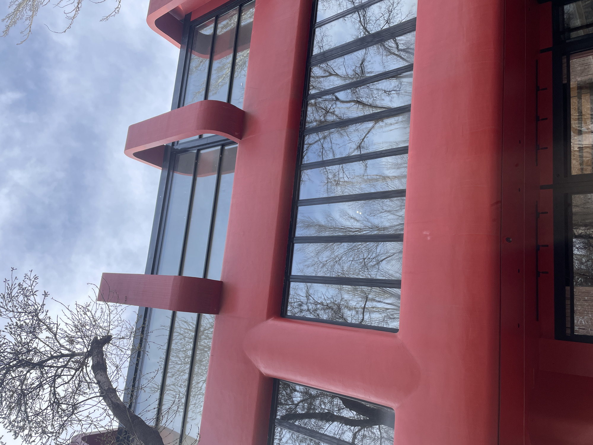 The image displays an upward-angled view of a section of the Cafesjian Center for the Arts in Yerevan, Armenia. The dominant architectural structure is a vibrant, matte red, characterized by smooth, curvilinear, and tubular forms. Two prominent, large, U-shaped or looped red structural elements are visible extending upwards from the top horizontal beam of the building. Below these, a thick horizontal red beam intersects with vertical segments, forming a pronounced 'T' shape. The building's facade primarily consists of multiple large, rectangular glass windows with black frames, which are interspersed between the red structural components. These windows extensively reflect bare deciduous tree branches and a cloudy, light blue sky. A section of a large, leafless tree is visible in the upper left background, its branches silhouetted against the sky. No individuals, specific actions, or interactions are present within the frame.
