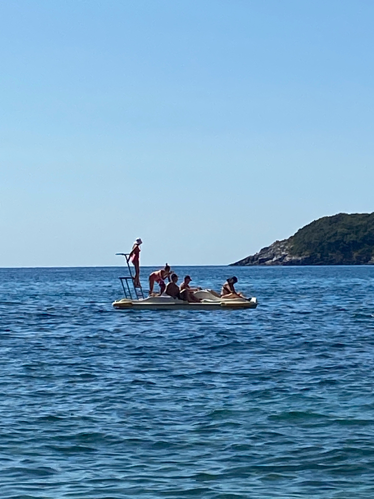 An aerial view shows a clear blue sky over a vast expanse of calm blue water. In the middle ground, a yellow pedal boat carries five people. One person, wearing a pink top and a white hat, stands on a raised platform at the front of the boat, holding onto a railing. Four other individuals are seated in the boat. In the background, a verdant, tree-covered hill rises from the water, indicating the presence of land, likely part of the coastline near Prijevor, Montenegro. The water exhibits gentle ripples, reflecting the bright sunlight.