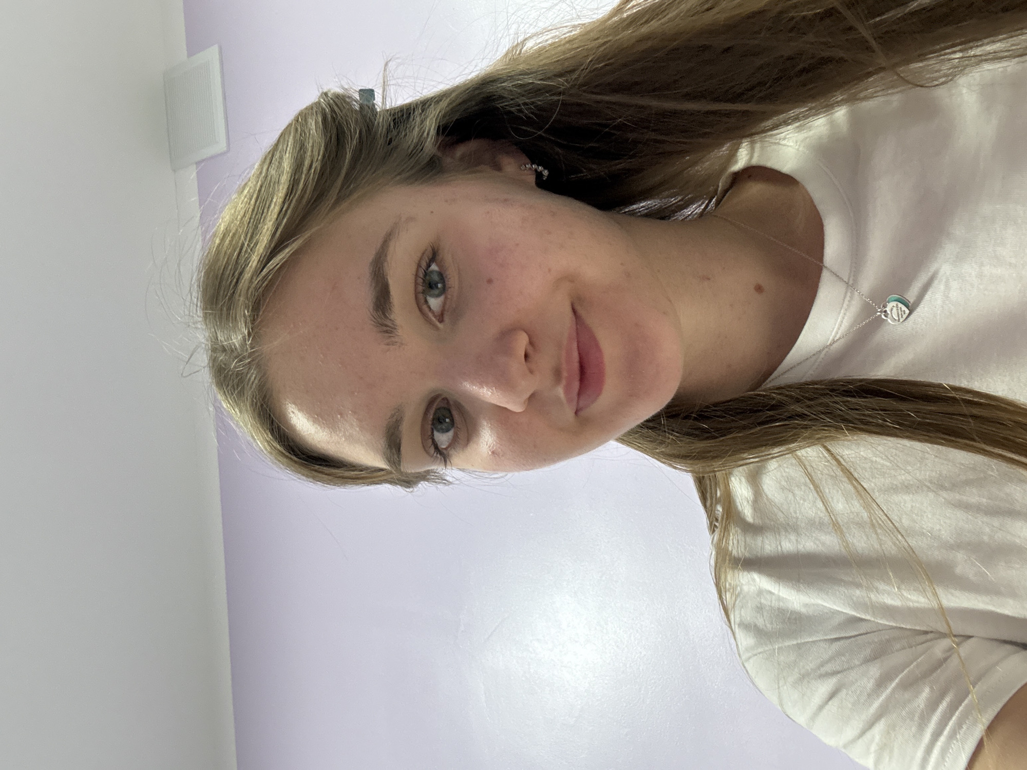 A young woman with long, light brown hair is centered in the image. She is wearing a white t-shirt and a silver necklace with a blue pendant. Her eyes are blue-green. The background consists of a light lavender wall with a white rectangular vent near the top left corner. The location is not discernible as Perm, Russia.